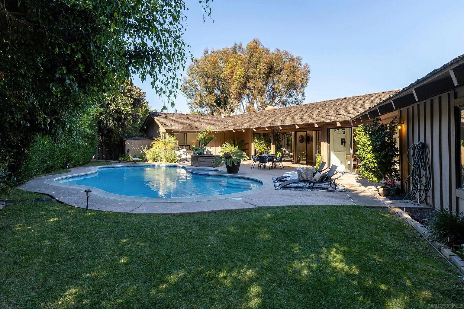 This image showcases a beautiful backyard featuring a kidney-shaped pool surrounded by a concrete patio and lush green lawn. The house, with its wood siding and large windows, provides a scenic backdrop. Outdoor furniture, including lounge chairs and a dining set, suggests a relaxing and inviting space for entertaining.