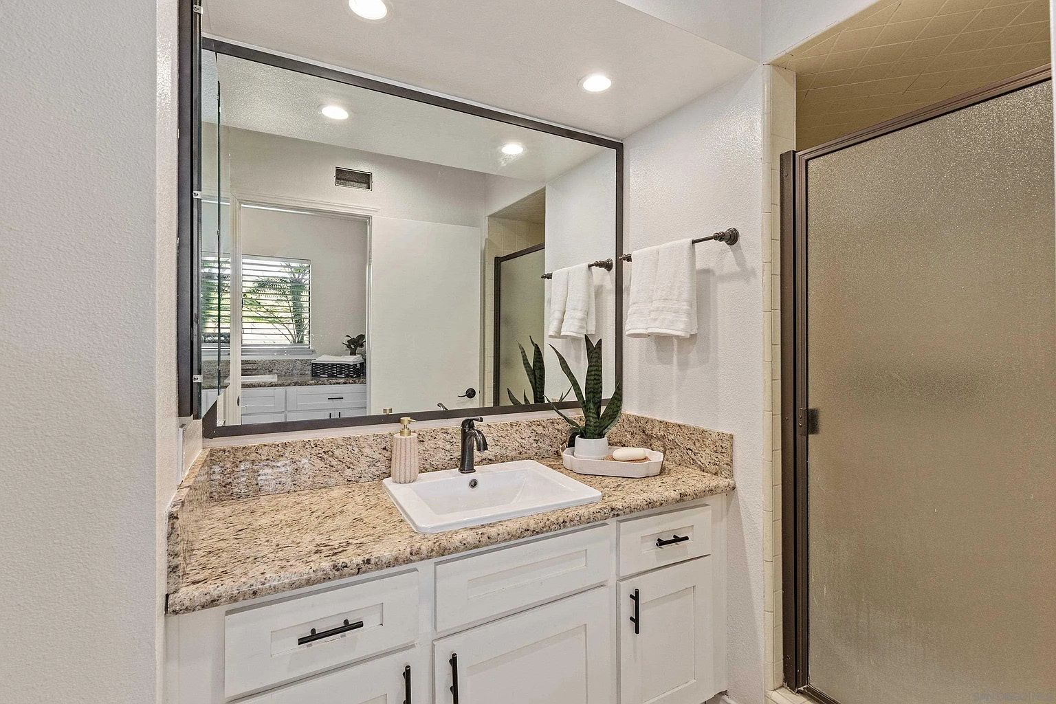 This is a well-lit bathroom featuring a granite countertop vanity with white cabinets and black hardware. A large mirror reflects the space, including a glimpse into another room. A glass-enclosed shower is visible to the right, and the overall impression is clean and functional.