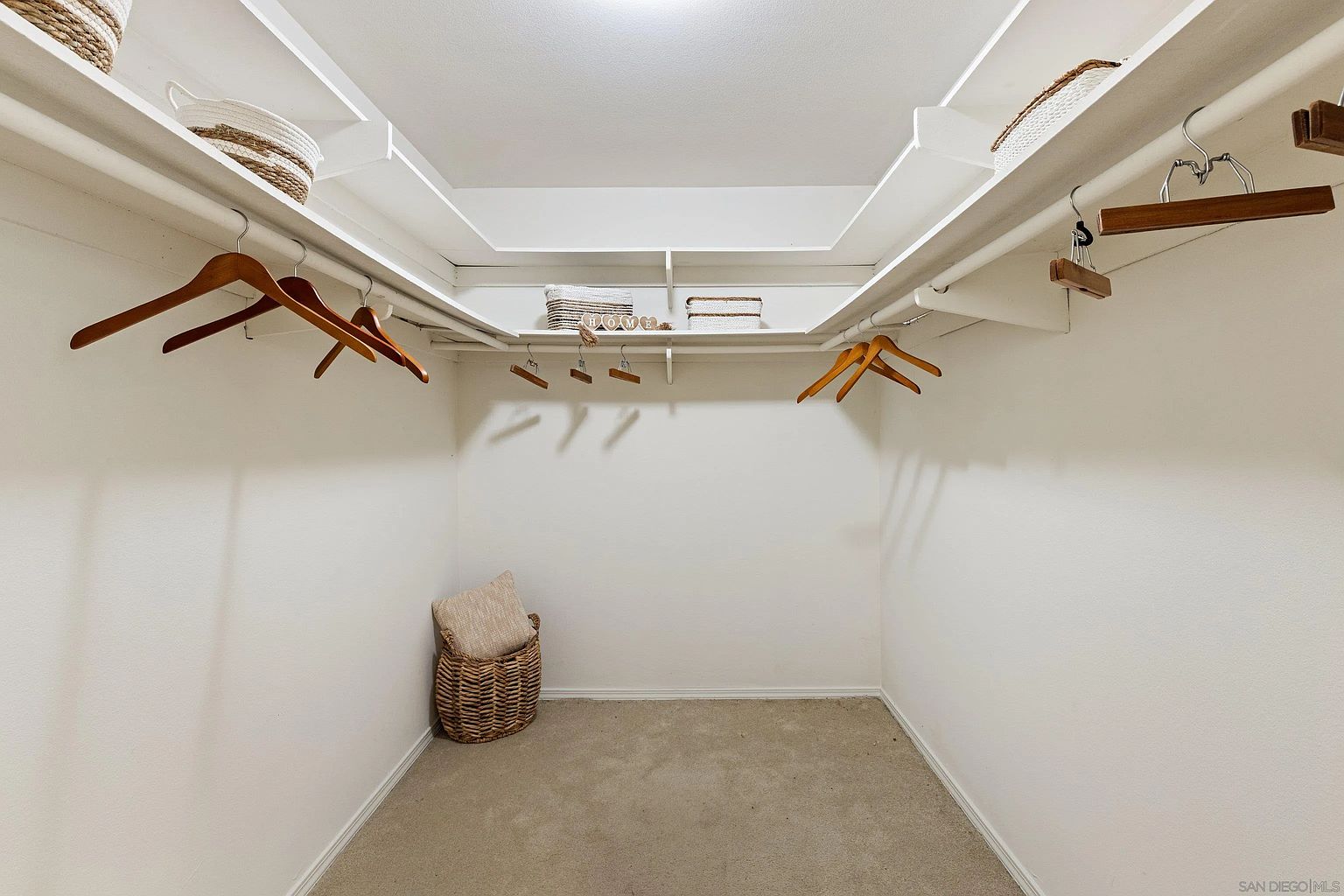 This is a well-organized walk-in closet with white walls, shelving, and hanging rods. Baskets and wooden hangers are used for storage, creating a clean and functional space. The closet is carpeted and appears spacious, offering ample storage for clothing and accessories.