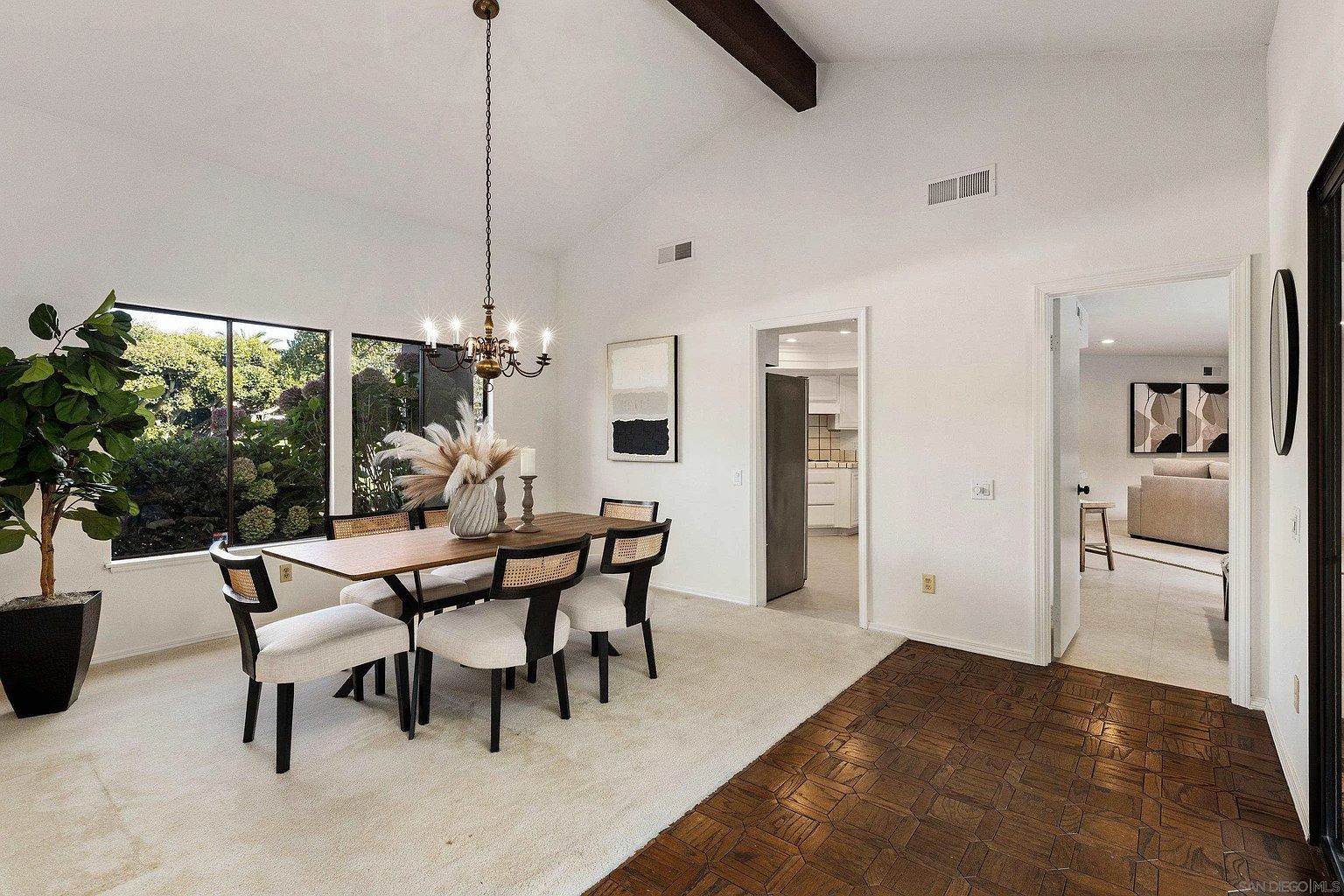 This is an interior shot of a dining room featuring a wooden table with seating for six, complemented by a chandelier overhead. Large windows provide natural light and a view of the greenery outside. The room is decorated in a neutral color palette with white walls and a light-colored carpet, creating a bright and inviting atmosphere.