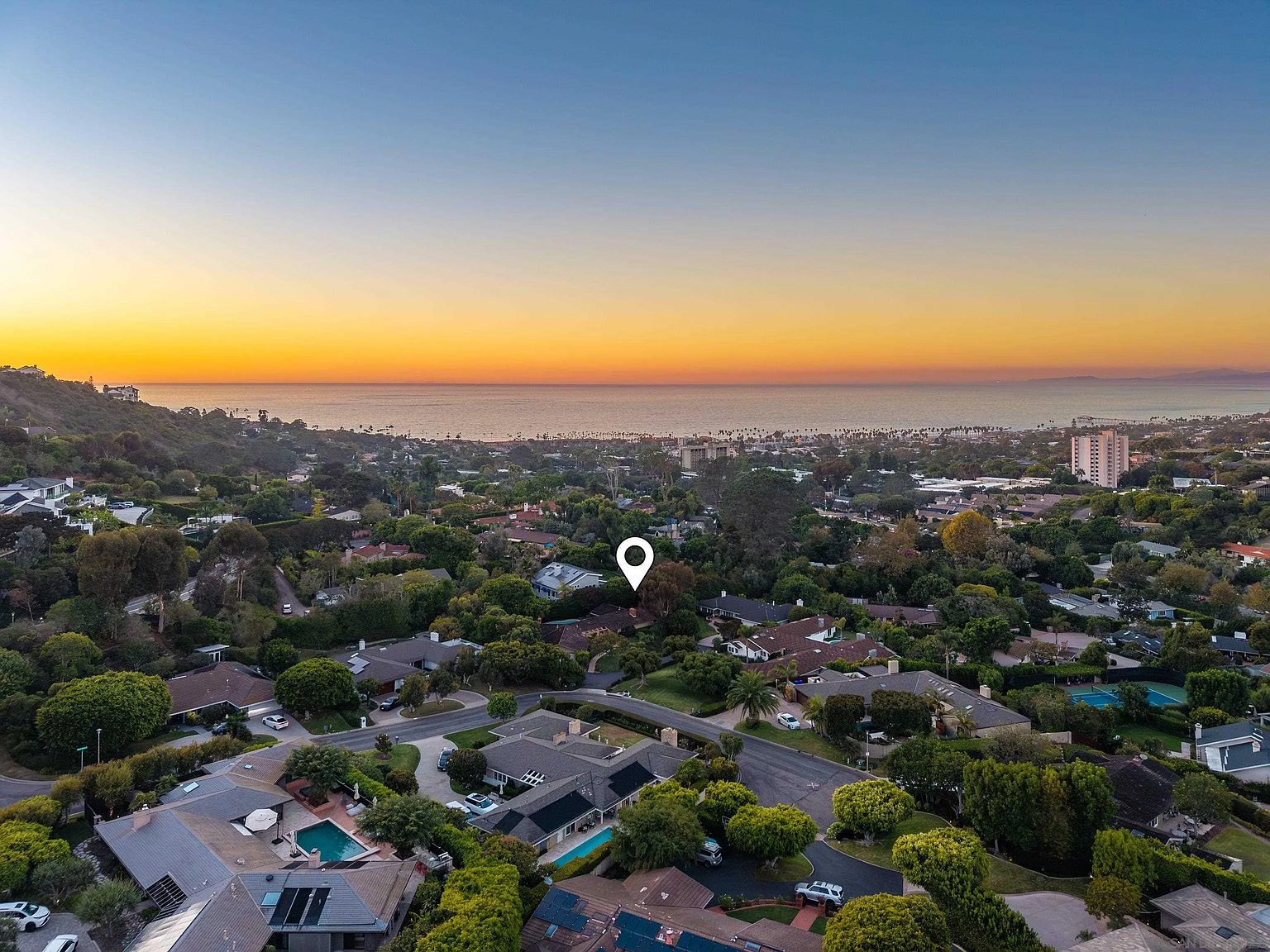 This aerial view showcases a residential neighborhood with lush greenery and mature trees, offering a sense of privacy and tranquility. Several homes feature pools and well-maintained yards, suggesting a desirable and upscale community. The ocean is visible in the background, adding to the property's appeal and highlighting its proximity to coastal amenities.