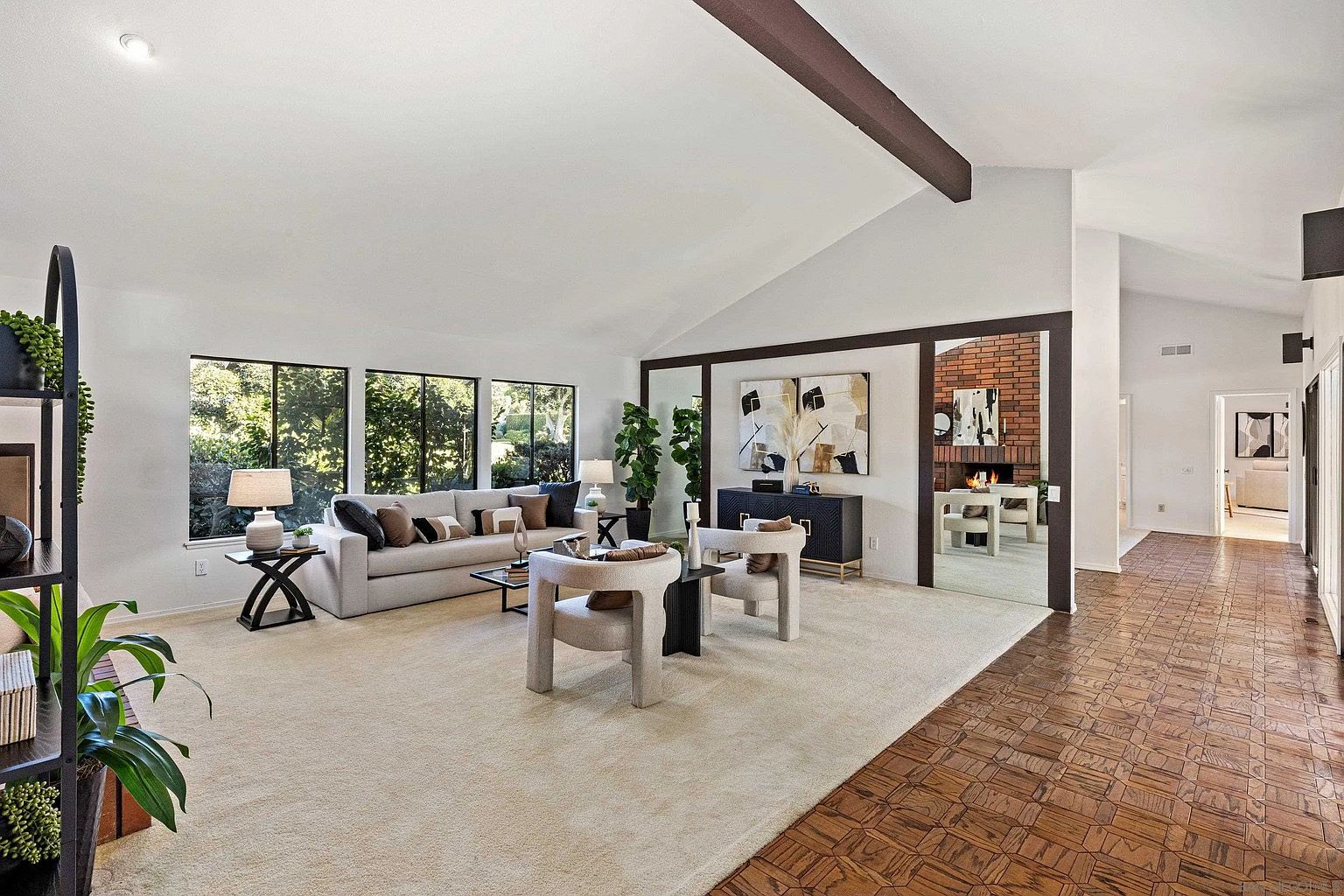 This is an interior shot of a living room featuring a neutral color palette with white walls and beige carpeting. The room is well-lit by natural light streaming through large windows overlooking greenery. A light-colored sofa is adorned with decorative pillows, and two modern chairs face the camera, creating an inviting and stylish space.