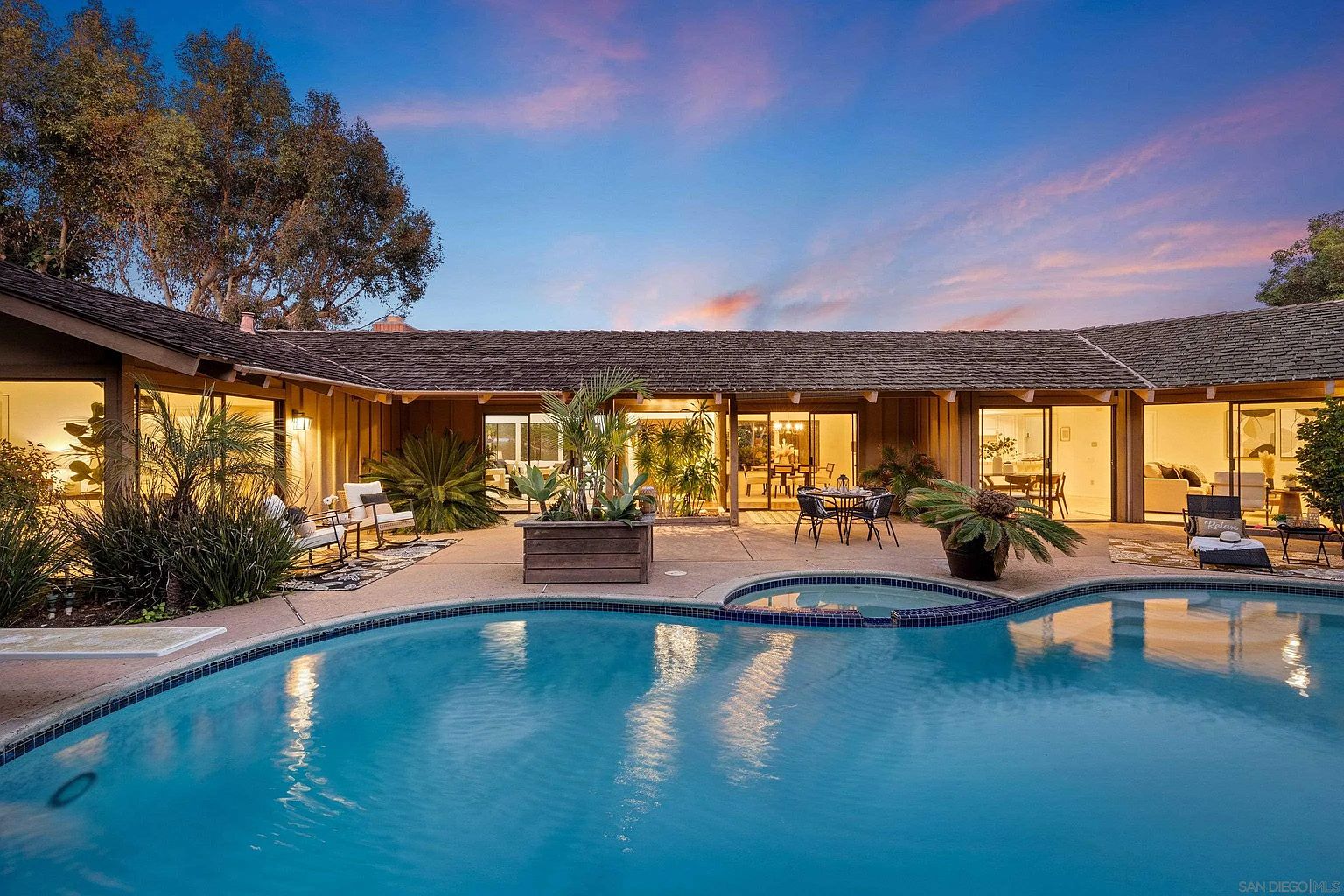 This image showcases a luxurious backyard featuring a large, inviting pool with a built-in spa. The pool is surrounded by a spacious patio area, complete with outdoor seating and lush landscaping. The single-story home provides a beautiful backdrop, with multiple glass doors offering seamless indoor-outdoor living. The perspective is from the pool looking towards the house.