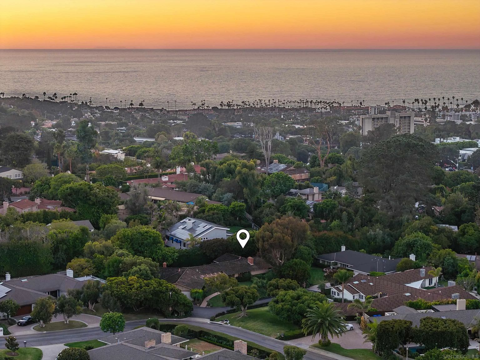 This aerial view showcases a neighborhood with lush greenery and mature trees, offering a sense of privacy and tranquility. The homes feature a variety of architectural styles, with glimpses of rooftops and well-maintained yards. In the background, the ocean stretches to the horizon under a warm, golden sky, highlighting the desirable coastal location.