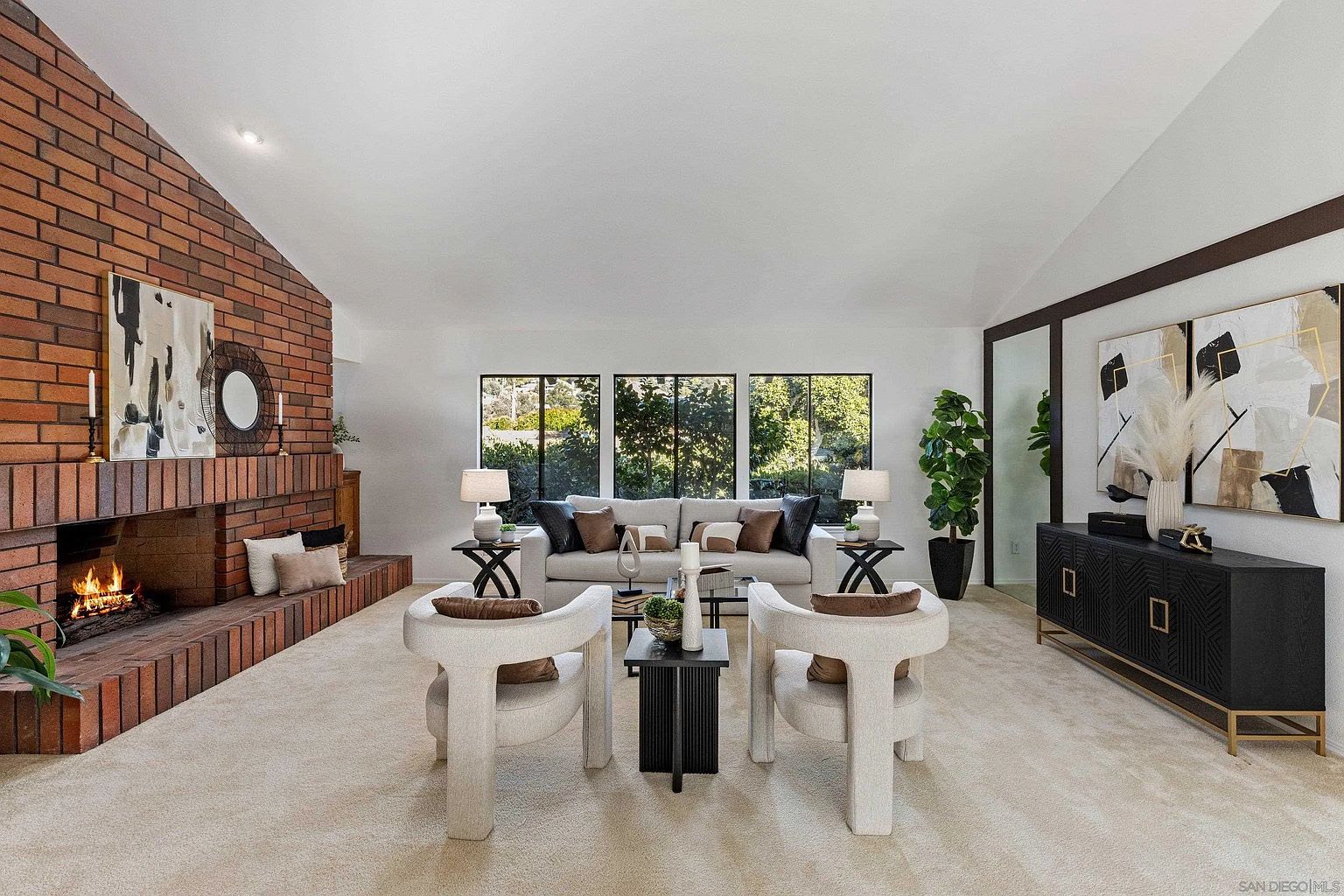 This is a bright and spacious living room featuring a brick fireplace, neutral-toned furniture, and large windows that provide ample natural light. The room is decorated with modern art and accessories, creating a stylish and inviting atmosphere. The perspective is from the front of the room, showcasing the layout and design elements.