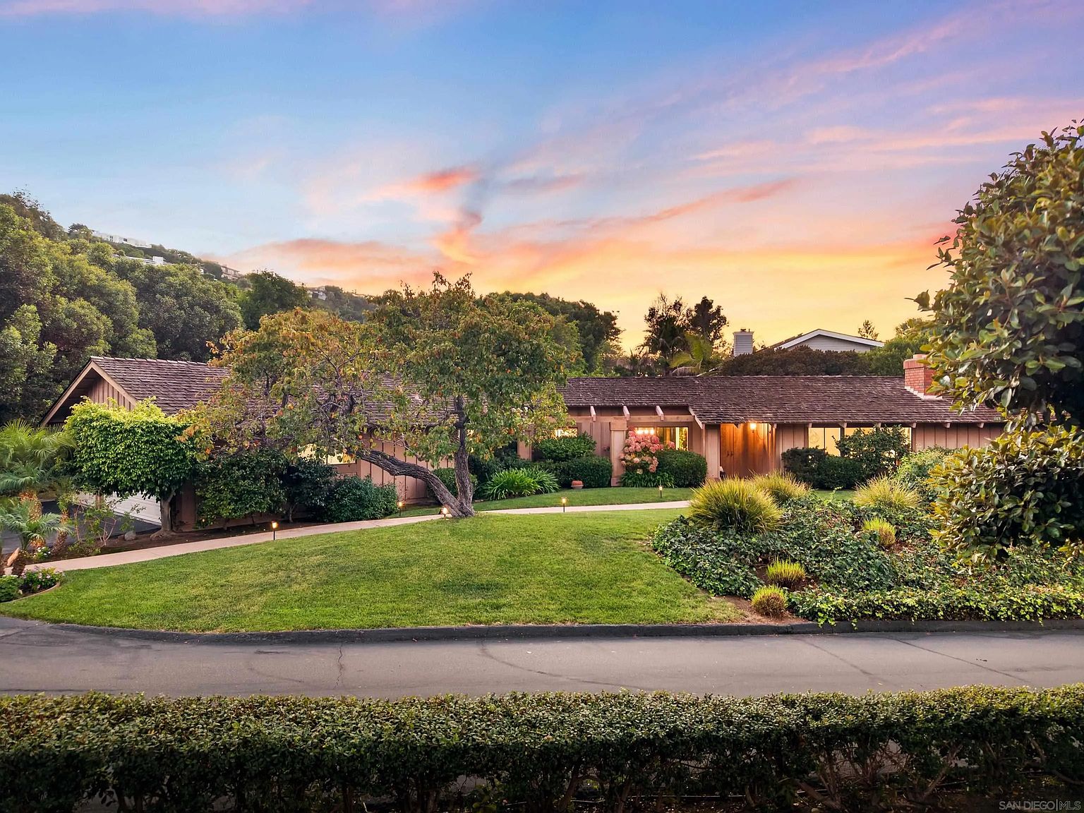 This image showcases the front exterior of a single-story home with a well-manicured lawn and mature landscaping. The house features a wood exterior, a low-pitched roof, and large windows that suggest ample natural light. The setting sun casts a warm glow, highlighting the property's curb appeal and inviting atmosphere.