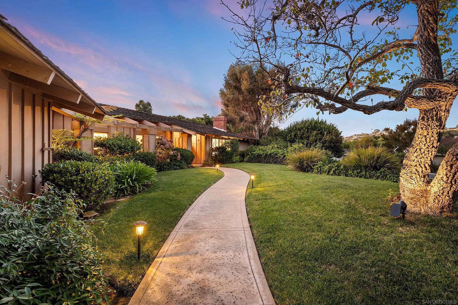 This image showcases a beautifully landscaped front yard with a well-maintained lawn and a winding concrete pathway leading to the house. The pathway is illuminated by landscape lighting, creating a warm and inviting ambiance. Mature trees and lush shrubbery add to the property's curb appeal, suggesting a serene and private setting.