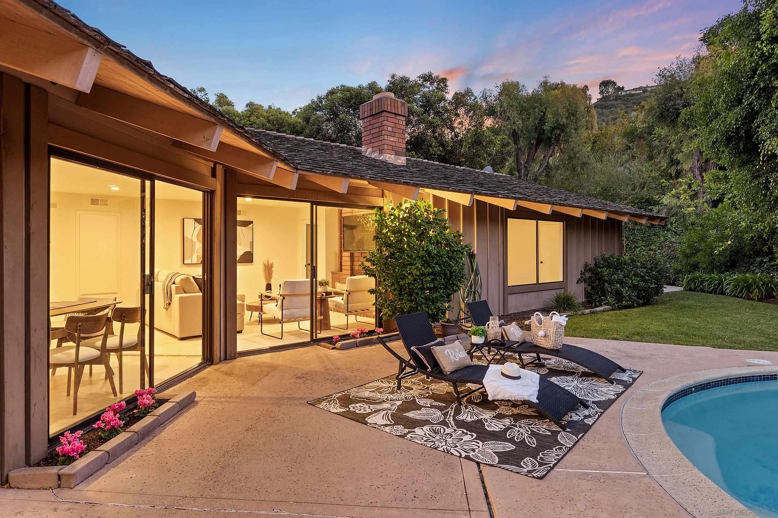 This is a rear exterior view of a home featuring a patio area with lounge chairs and a pool. Large sliding glass doors provide access to the interior, showcasing a dining area and living space. The backyard is well-maintained with lush greenery, creating a private and relaxing atmosphere.