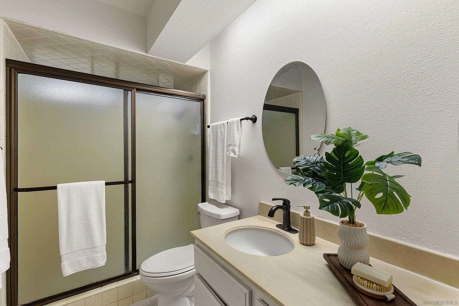This is a well-lit bathroom featuring a vanity with a light-colored countertop and white cabinets. An oval mirror hangs above the sink, complemented by a modern black faucet and decorative soap dispenser. A shower with a frosted glass door is visible to the left, and a toilet is positioned next to the vanity, creating a functional and clean space.