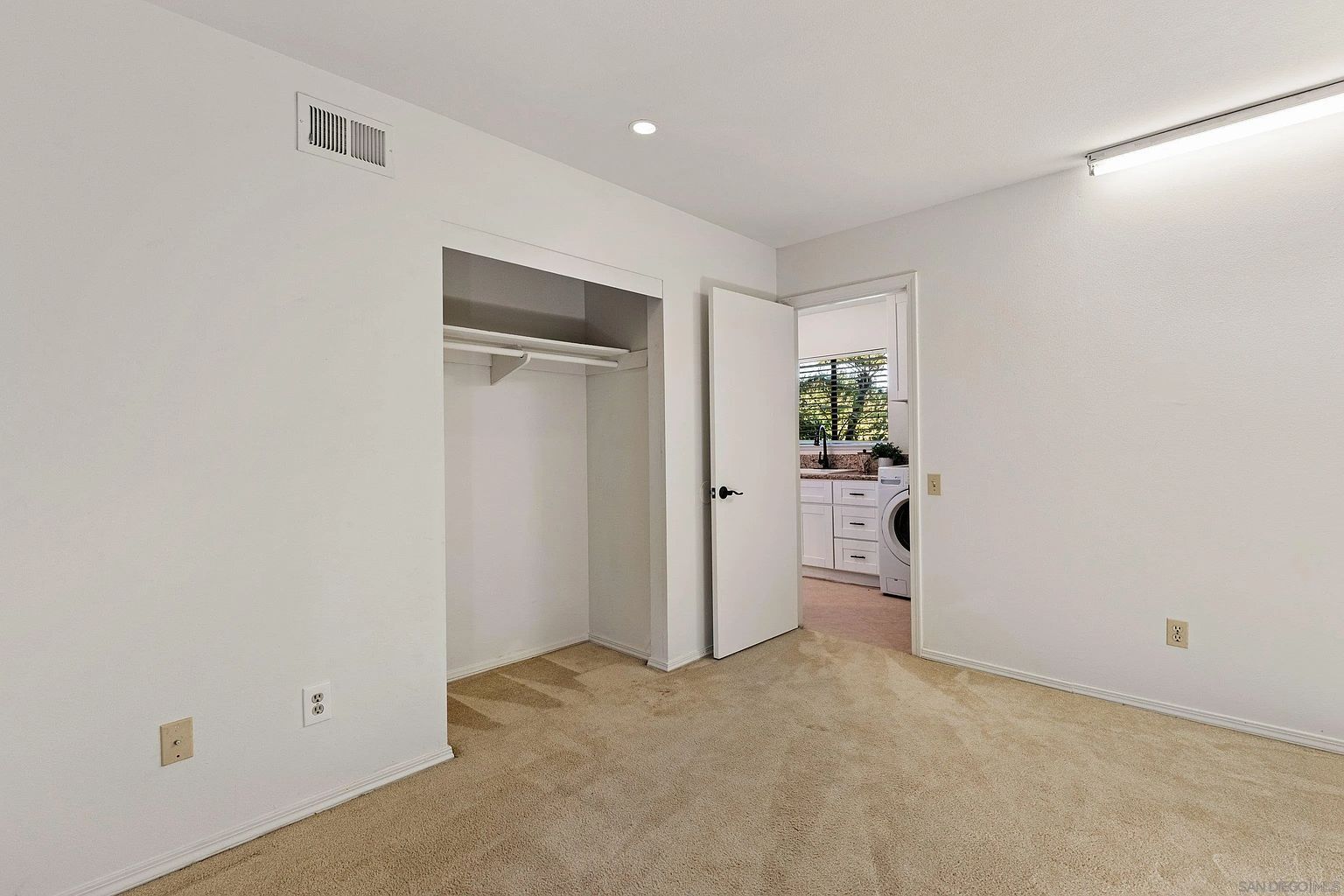 The image showcases a bright, empty bedroom with neutral beige carpeting and white walls. A closet with a hanging rod is visible, and a doorway leads to what appears to be a laundry area with a washing machine and a glimpse of a kitchen. The room is well-lit, creating a clean and inviting atmosphere.