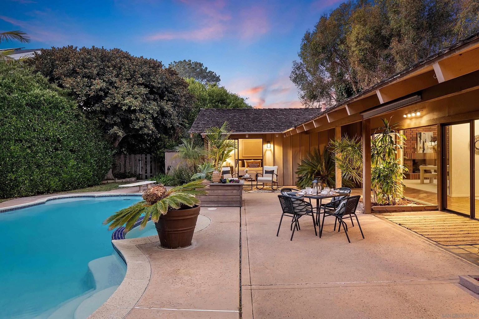 This image showcases a beautifully designed outdoor patio area, perfect for entertaining or relaxing. The space features a dining set, comfortable lounge seating, and lush greenery, creating a serene and inviting atmosphere. A swimming pool is visible in the background, adding to the appeal of this outdoor oasis.