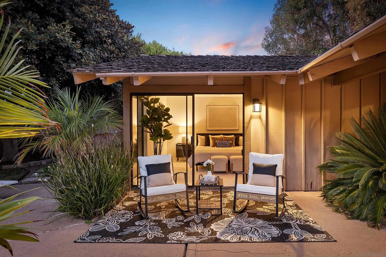 This image showcases a cozy patio area adjacent to a bedroom, creating an inviting indoor-outdoor living space. The patio features two rocking chairs with cushions, a small table, and a decorative rug, all set against the backdrop of a sliding glass door that reveals a glimpse of the bedroom interior. Lush greenery surrounds the patio, enhancing its tranquil and private atmosphere.