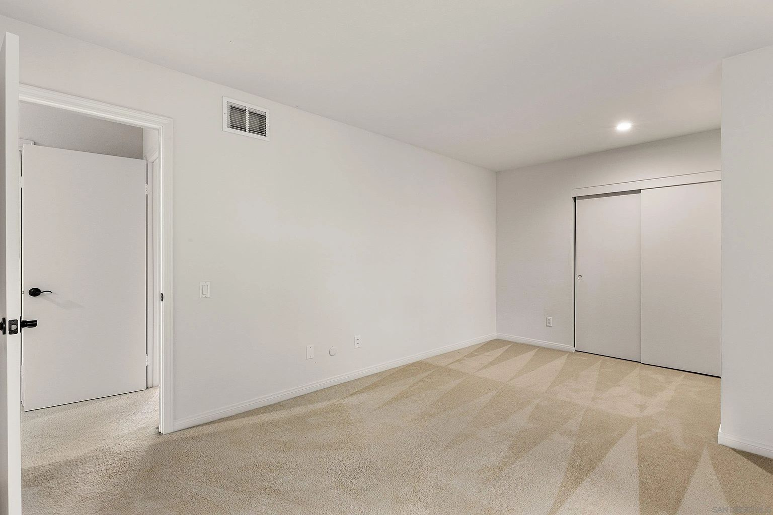 The image showcases a well-lit bedroom with neutral beige carpet and white walls. A closet with sliding doors is visible on the right, and a doorway leads to another room on the left. The room appears clean and spacious, offering a blank canvas for potential buyers to envision their own style.