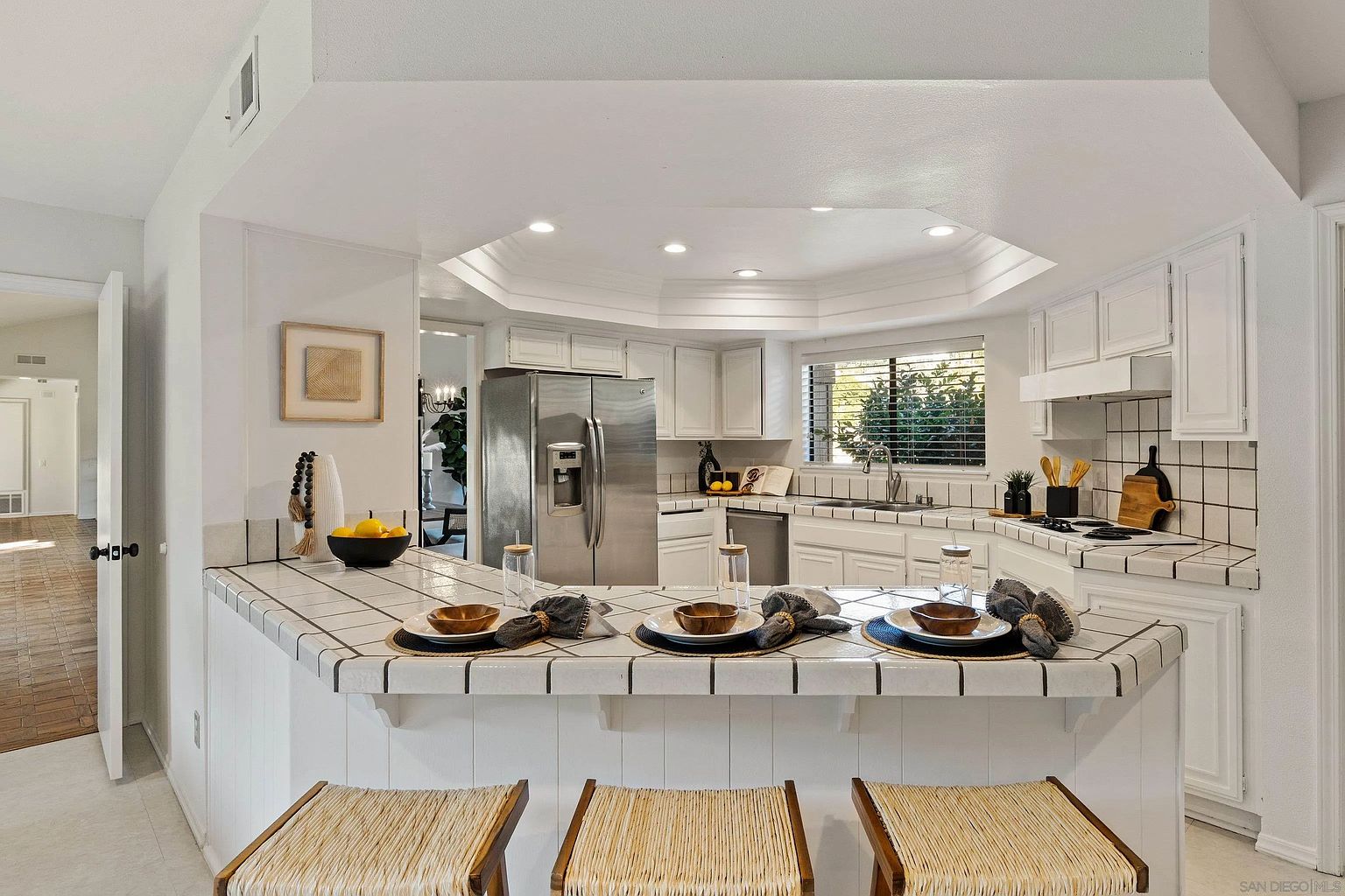 This is a bright and airy kitchen featuring white cabinetry, stainless steel appliances, and a tiled countertop with bar seating. The kitchen is well-lit with recessed lighting and natural light from a window above the sink. The overall impression is clean, inviting, and functional, perfect for a modern home.