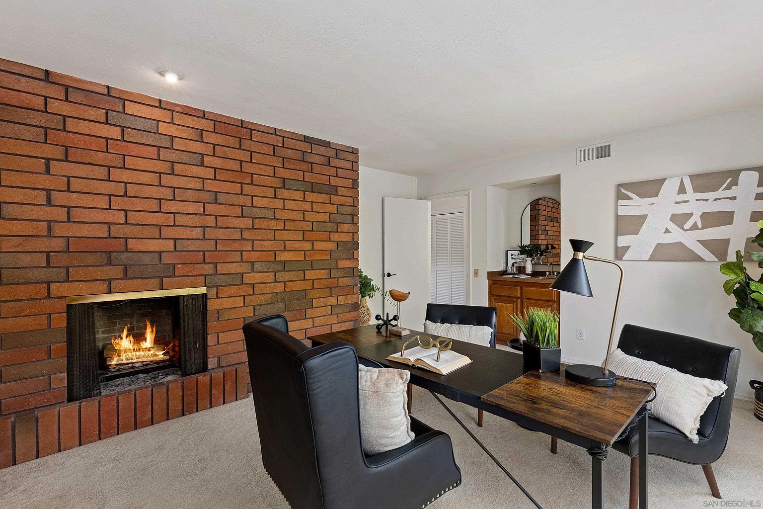 This is an interior shot of a home office featuring a brick fireplace. The room is furnished with a large desk, black leather chairs, and decorative pillows. A lamp illuminates the desk, and artwork hangs on the wall, creating a cozy and productive workspace.