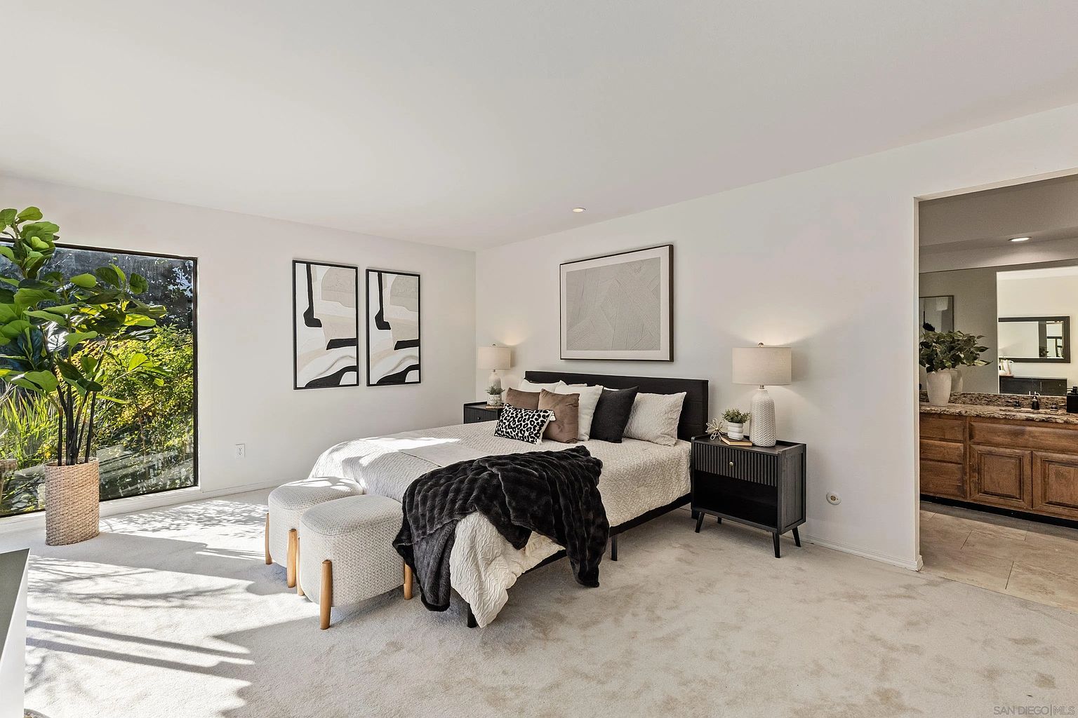 This is a well-lit primary bedroom featuring a bed with a dark headboard, adorned with multiple pillows and a dark throw blanket. Two upholstered ottomans are placed at the foot of the bed. The room has white walls, carpeted floors, and is decorated with framed artwork. A glimpse into an adjacent bathroom is visible through a doorway.