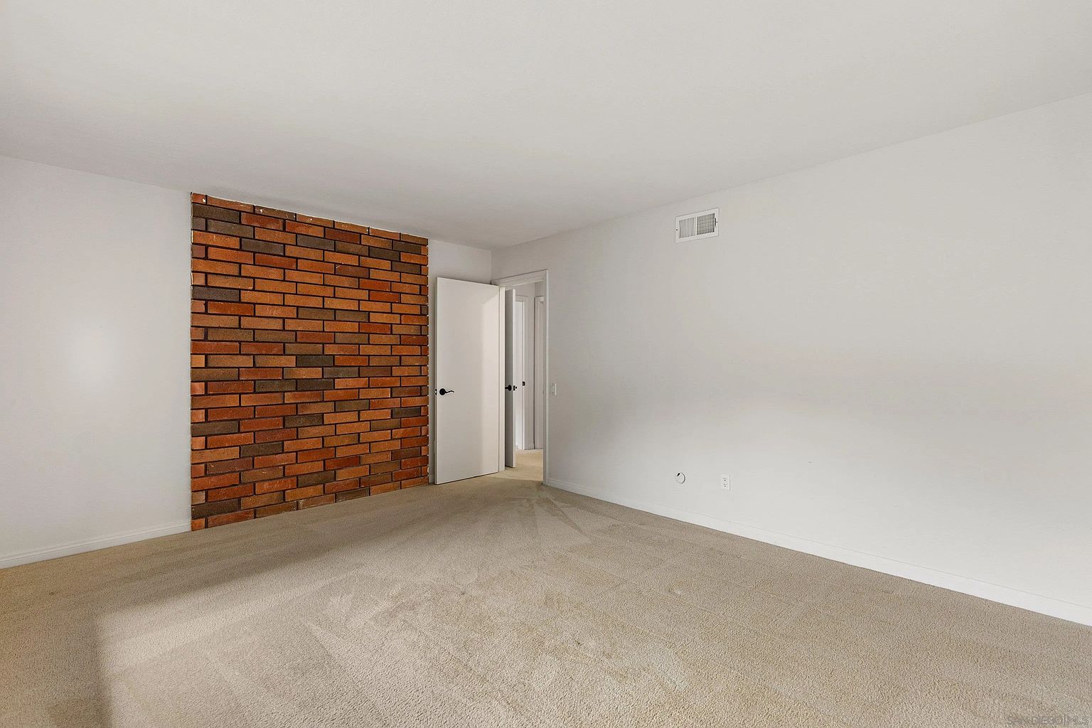 This is an interior shot of a bedroom featuring neutral-toned carpet and white walls, with a brick accent wall adding a touch of character. A doorway leads to another room, suggesting an en-suite or hallway. The room appears spacious and well-lit, offering a blank canvas for potential buyers.