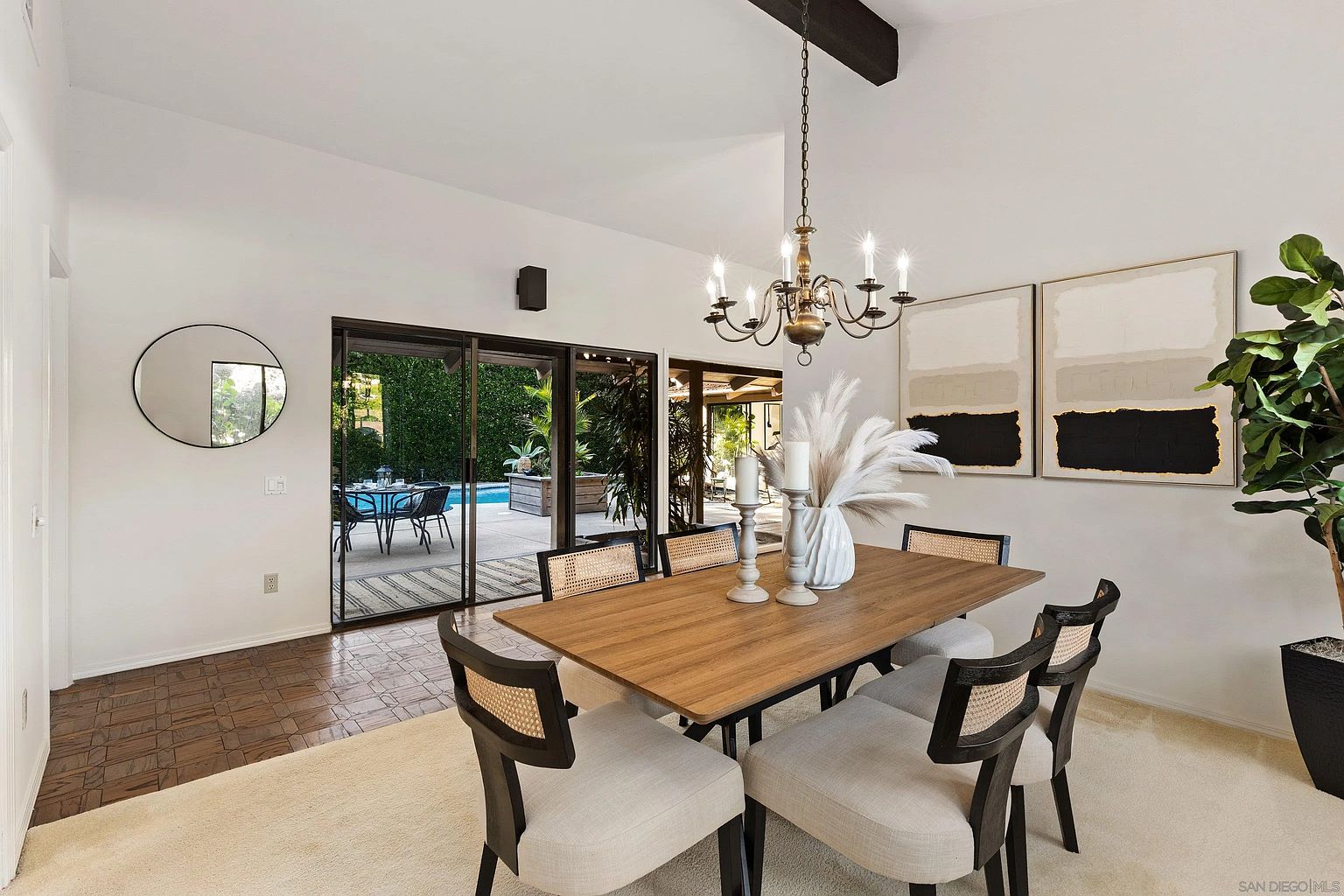 This is an interior shot of a dining room featuring a wooden table with six chairs, a chandelier, and abstract art on the wall. Sliding glass doors offer a view of the outdoor pool area, enhancing the room's connection to the exterior. The room is well-lit and styled with modern decor, creating an inviting and sophisticated atmosphere.