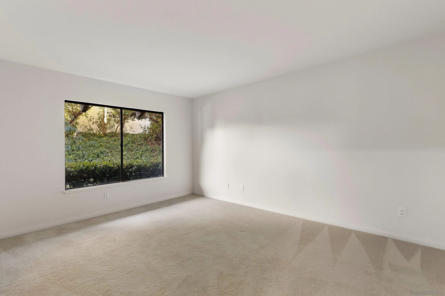 This is a bright and airy bedroom with neutral beige carpet and white walls. A large window provides natural light and a view of lush greenery outside. The room is simple and clean, offering a blank canvas for potential buyers to envision their own style.