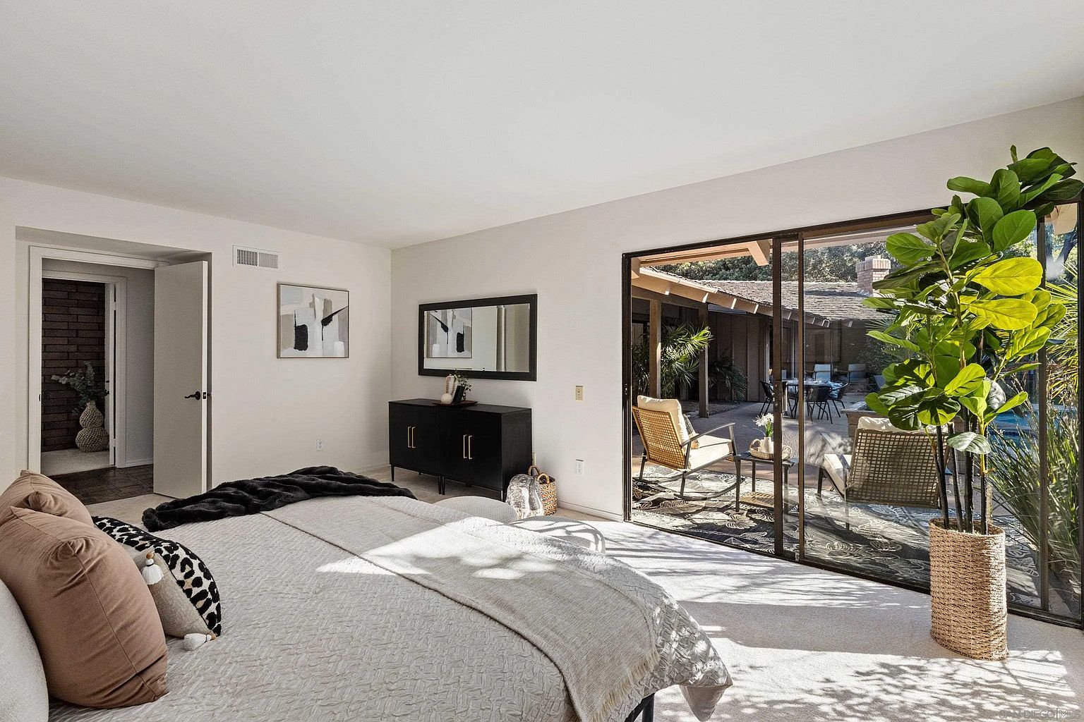 This is a bright primary bedroom featuring a large sliding glass door that opens to an outdoor patio area. The room is decorated with neutral tones, including a light-colored bedspread and a black dresser with a mirror above it. A large potted plant sits near the sliding door, adding a touch of nature to the space.
