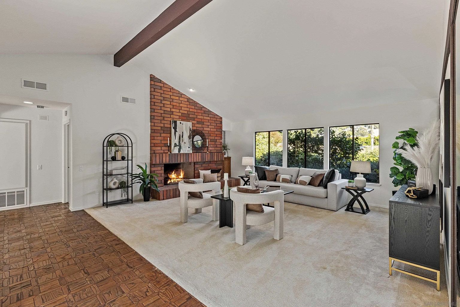 This is an interior shot of a living room featuring a brick fireplace, a light-colored sofa with decorative pillows, and two modern armchairs. The room has a high ceiling with a wooden beam, large windows providing natural light, and a neutral-toned carpet. The overall impression is a cozy and stylish living space.