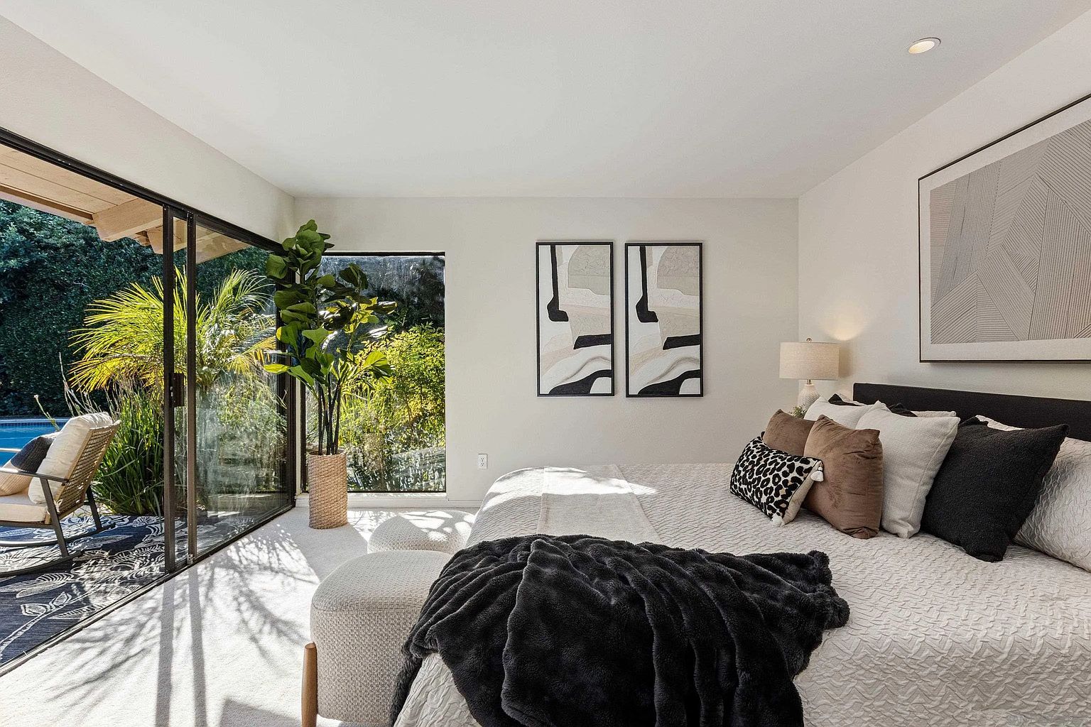 This is a bright and airy primary bedroom featuring a large bed with multiple pillows and a dark throw blanket. The room has a modern aesthetic with neutral tones, complemented by abstract artwork on the walls. A sliding glass door provides access to an outdoor patio and lush greenery, enhancing the connection to nature.