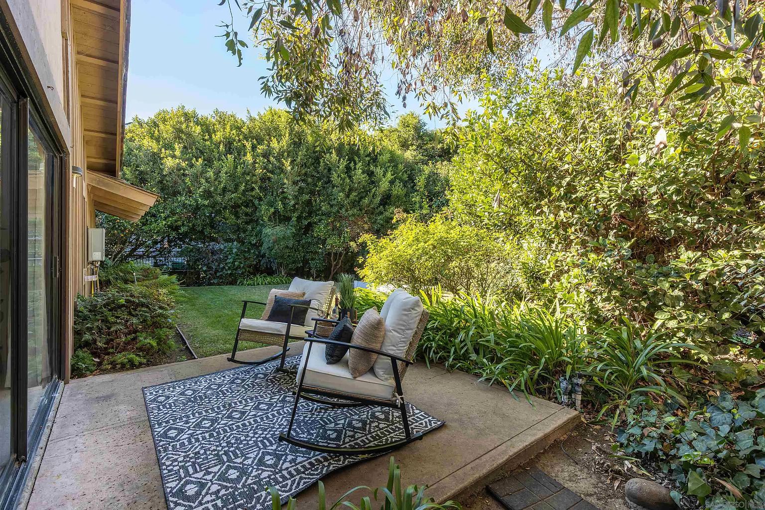 This image showcases a cozy patio area with two rocking chairs placed on a patterned outdoor rug. The patio is surrounded by lush greenery, including trees and various plants, creating a private and serene outdoor living space. The setting suggests a relaxing and inviting atmosphere, perfect for enjoying the natural surroundings.