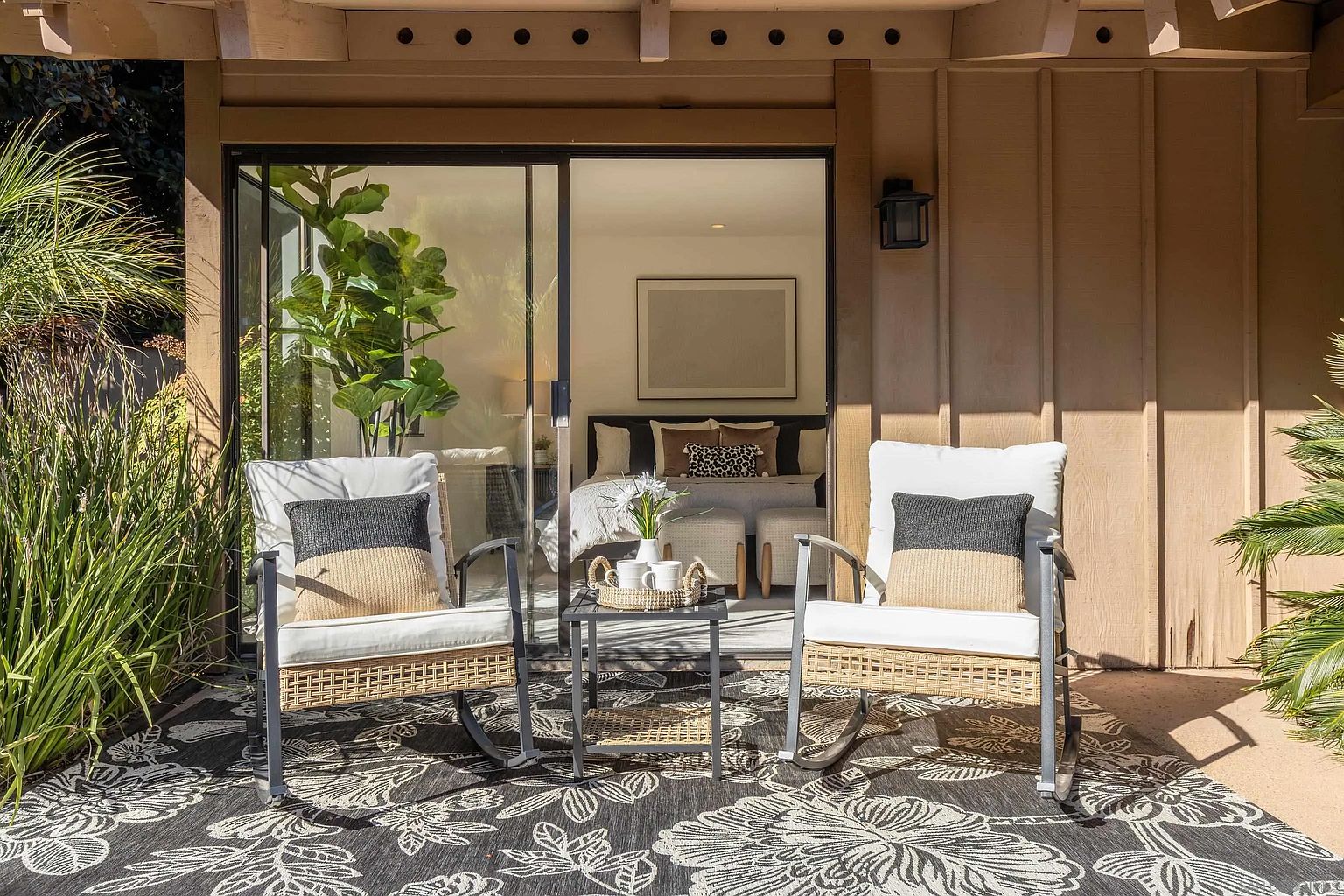 This image showcases a cozy and inviting outdoor patio area. Two wicker rocking chairs with white cushions and decorative pillows flank a small table set with mugs, creating a relaxing seating arrangement. The patio is adorned with a floral-patterned rug, and a sliding glass door provides access to the interior, offering a seamless transition between indoor and outdoor living spaces.