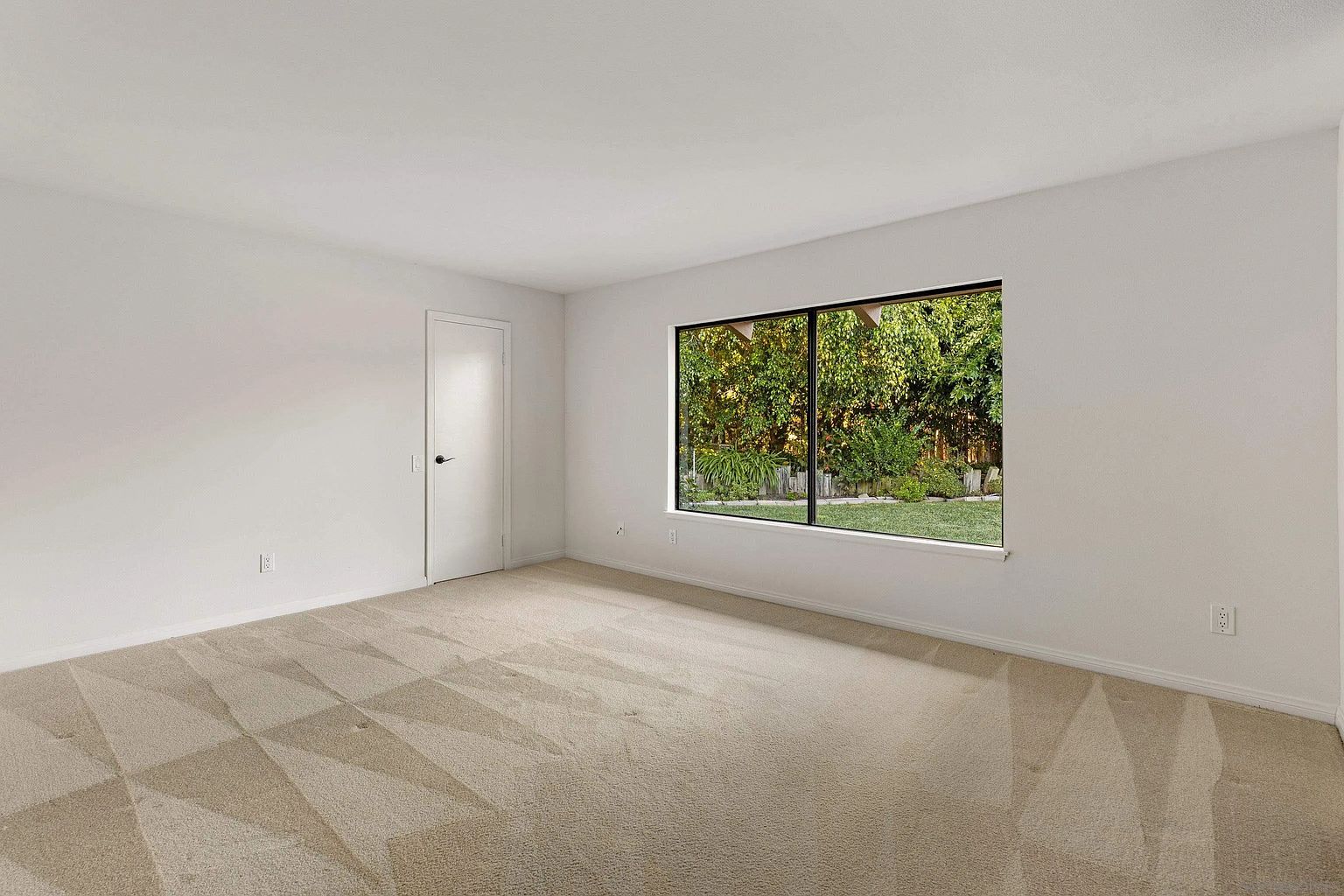 This is an interior shot of a bedroom, likely a guest or secondary bedroom. The room is painted in a neutral white tone, with a beige carpet featuring a subtle geometric pattern. A window provides a view of the exterior greenery, and a closed door is visible on the left side of the frame.