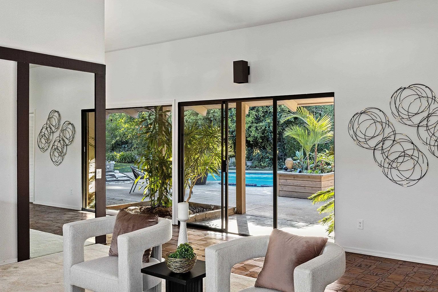 This interior shot showcases a modern living room with a view of the backyard pool area through sliding glass doors. Two light gray armchairs with mauve pillows flank a dark side table with a potted plant, creating a cozy seating arrangement. The room features unique wall art and a mix of flooring, including wood and carpet, adding to the contemporary aesthetic.