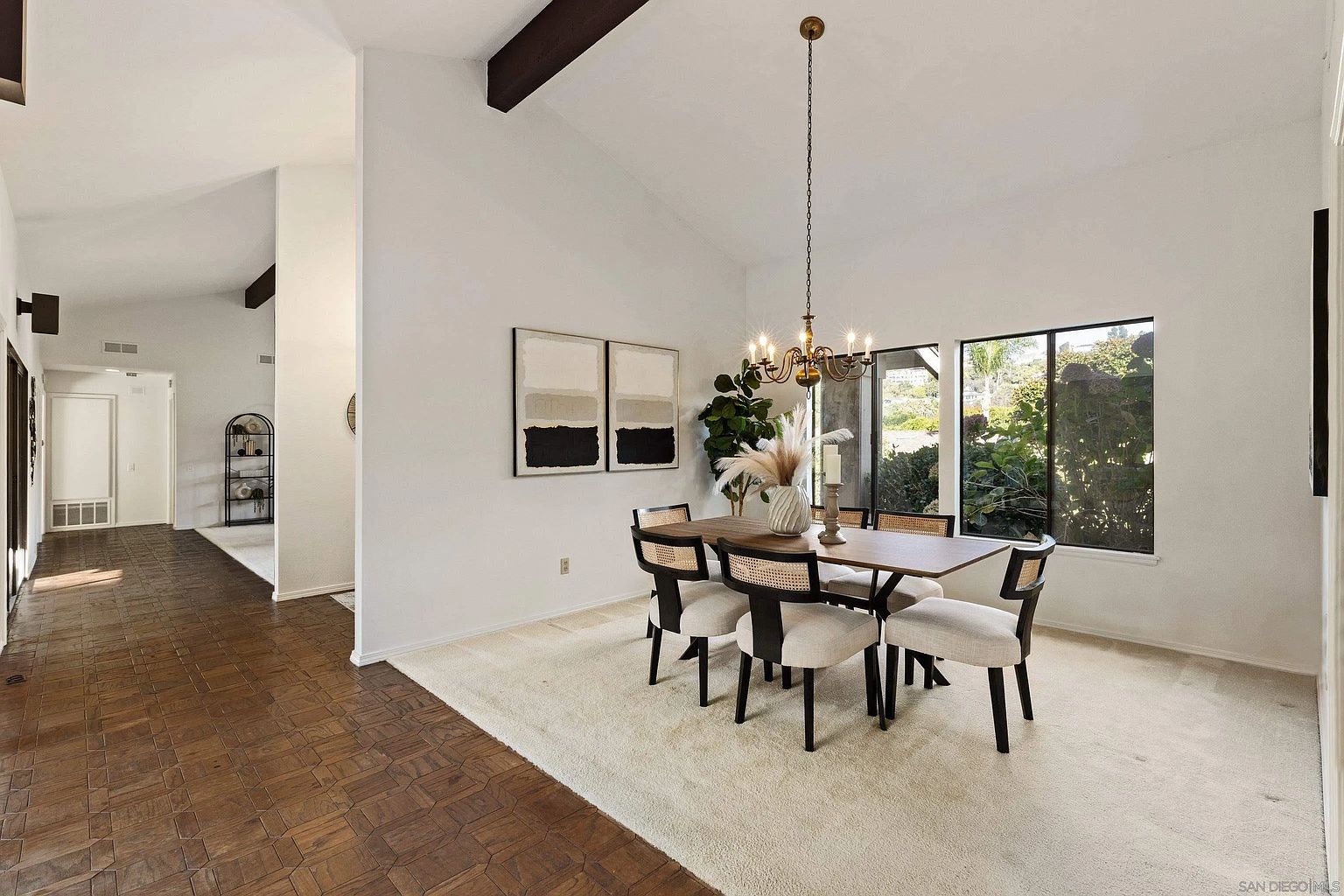 This is an interior shot of a dining room featuring a wooden dining table with four chairs, a chandelier hanging above, and two abstract paintings on the wall. The room is bright and airy, with natural light coming in through a window overlooking greenery. The overall impression is one of modern elegance and comfort.