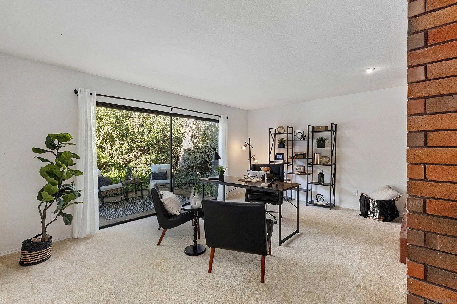 This is an interior shot of a well-lit home office. The room features a modern desk with a black frame and a dark wood top, complemented by two black chairs. A tall black bookshelf stands against the wall, and a large sliding glass door provides natural light and a view of the outdoor patio.
