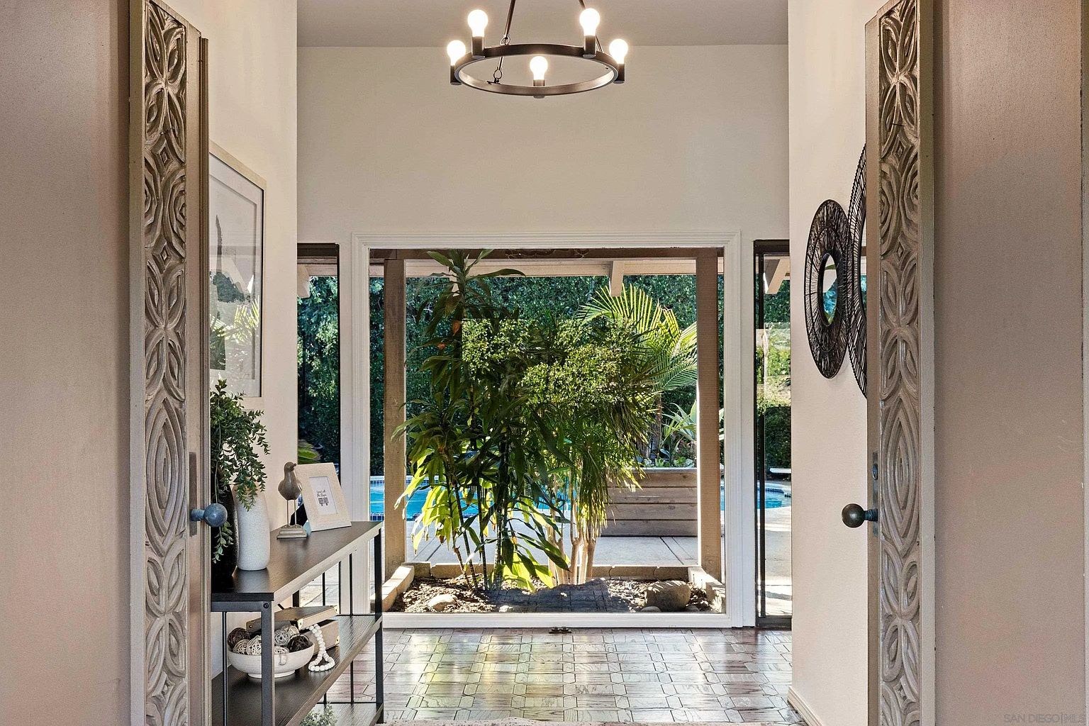 This interior shot showcases a hallway with a unique indoor garden visible through a large window, offering a glimpse of the pool area. The hallway features decorative doors, a stylish console table, and tiled flooring, creating an inviting and sophisticated atmosphere. A modern chandelier hangs from the ceiling, adding a touch of elegance.