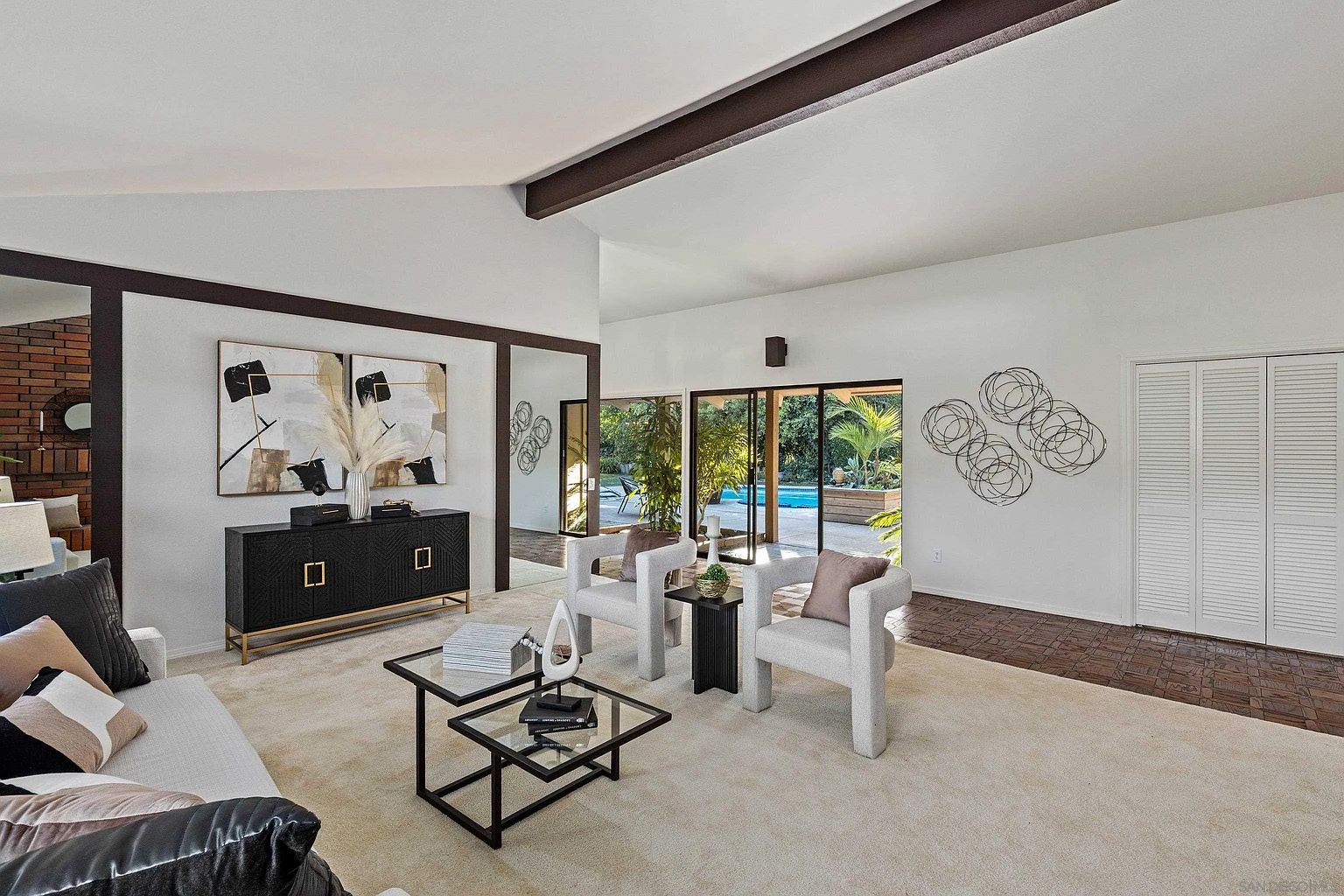 This is an interior shot of a living room featuring a modern design with a neutral color palette. The room includes two white armchairs, a glass coffee table, and a black sideboard with artwork above it. Large sliding glass doors provide a view of the outdoor pool area, enhancing the indoor-outdoor flow.