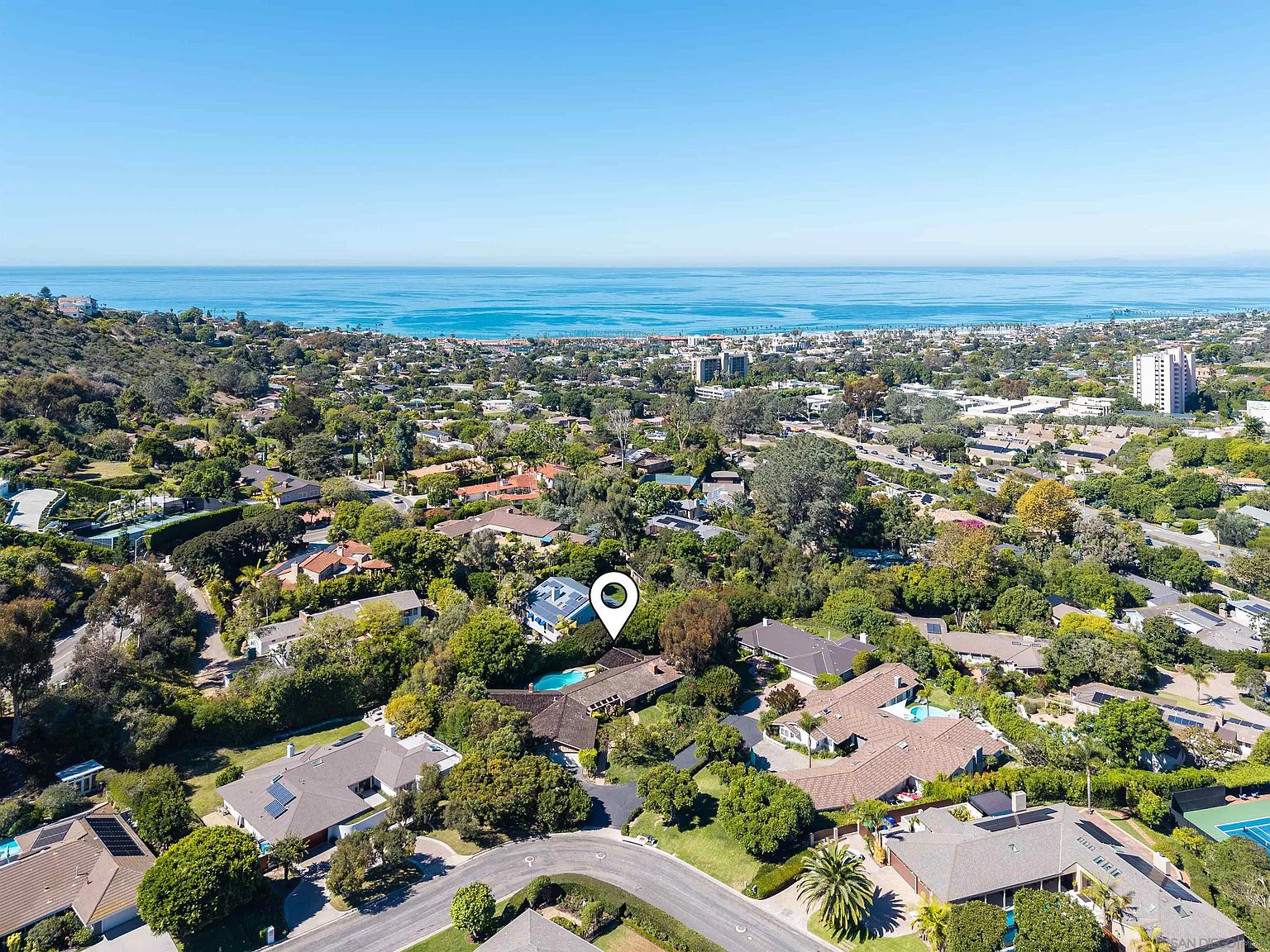 This aerial view showcases a luxurious residential area with lush greenery and stunning ocean views. The homes feature a variety of architectural styles, many with private pools and well-manicured gardens. The perspective highlights the desirable location and upscale neighborhood, emphasizing the proximity to both nature and urban amenities.