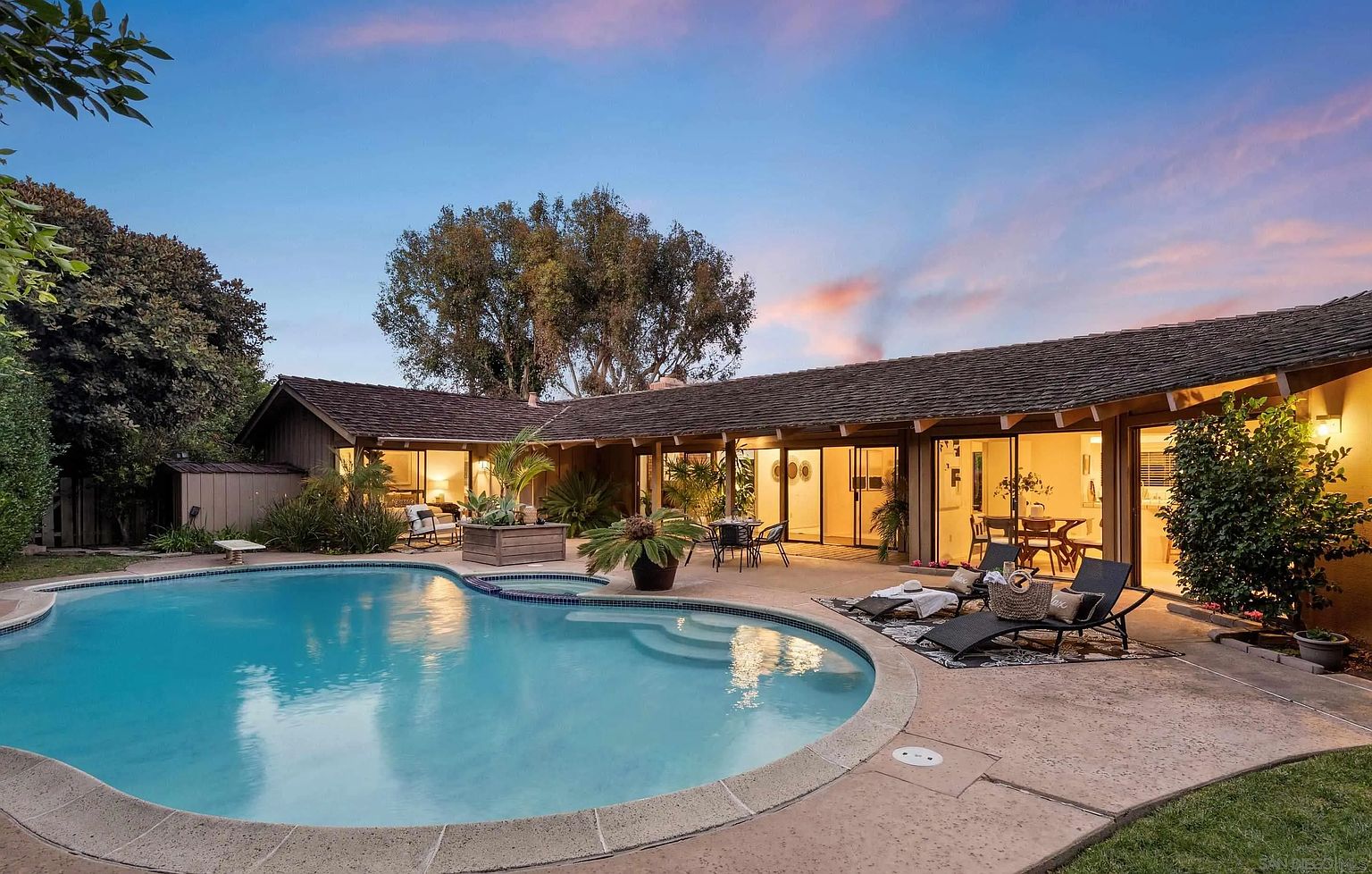 This image showcases a stunning backyard featuring a large, freeform pool with a built-in spa. The pool is surrounded by a spacious concrete patio, furnished with lounge chairs and outdoor seating. The house, with its expansive glass doors, seamlessly blends indoor and outdoor living, creating an inviting and luxurious atmosphere.