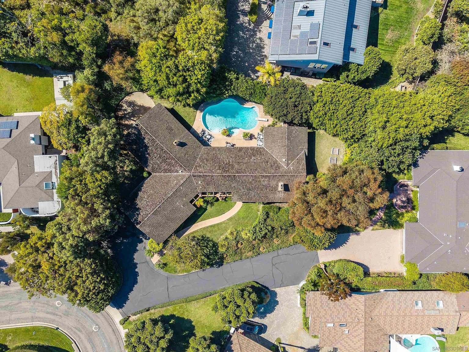 This aerial shot showcases a well-maintained property featuring a house with a unique roof design, a sparkling blue swimming pool, and lush green landscaping. A winding driveway leads to the house, surrounded by mature trees and manicured lawns. The image provides a comprehensive view of the property's layout and its integration with the surrounding environment.