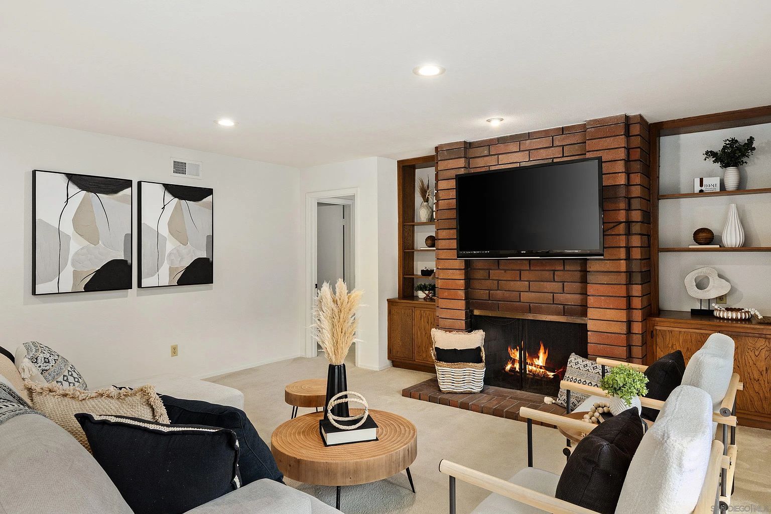 This is an interior shot of a living room featuring a brick fireplace with a mounted television. The room is decorated with modern art, stylish furniture, and decorative items, creating a cozy and inviting atmosphere. Built-in shelving flanks the fireplace, adding storage and display space.