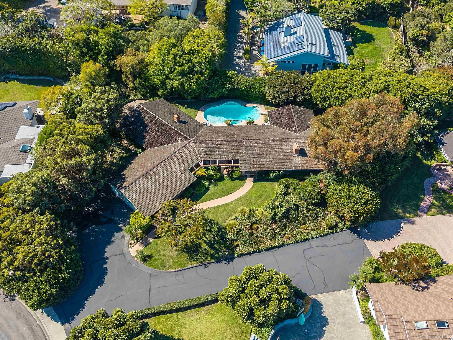 This aerial shot showcases a sprawling single-story home with a distinctive brown tile roof, nestled among lush greenery. A sparkling blue pool is visible in the backyard, adding to the property's appeal. The circular driveway and well-maintained landscaping enhance the curb appeal, creating an inviting and private estate.