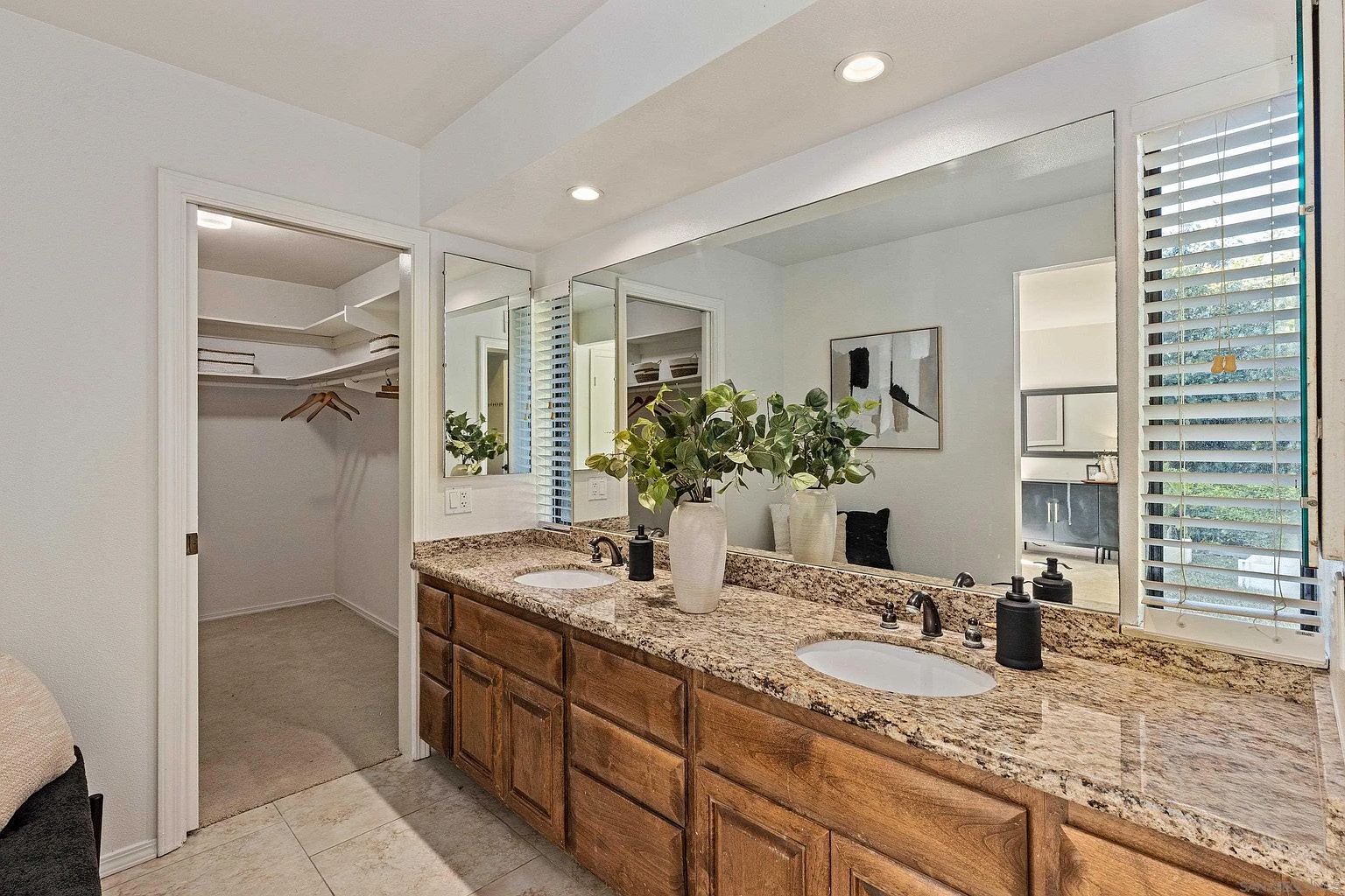 This is a well-lit primary bathroom featuring a double vanity with a granite countertop and wooden cabinets. Large mirrors extend across the wall above the sinks, reflecting natural light from the window with blinds. A walk-in closet is visible through an open doorway, adding to the bathroom's functionality and appeal.