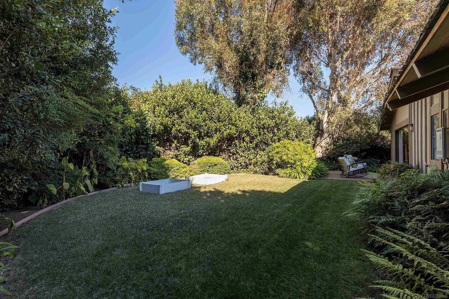 This is a view of a well-maintained backyard garden. The yard features a lush green lawn, mature trees, and various shrubs and plants, creating a serene and private outdoor space. A raised garden bed is visible, along with outdoor seating near the house, suggesting an area for relaxation and entertainment.