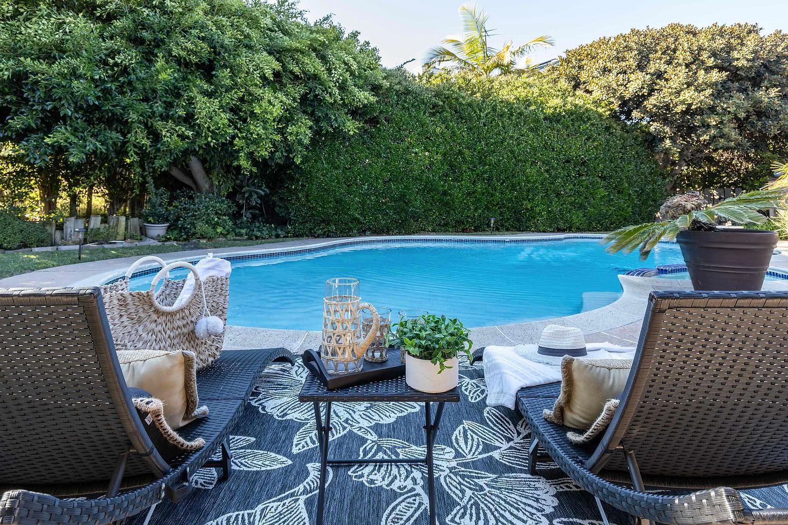 This image showcases a luxurious backyard pool area, complete with lounge chairs, a stylish outdoor rug, and a small table with decorative items. Lush greenery surrounds the pool, creating a private and serene atmosphere. The scene evokes relaxation and upscale outdoor living, perfect for entertaining or unwinding.