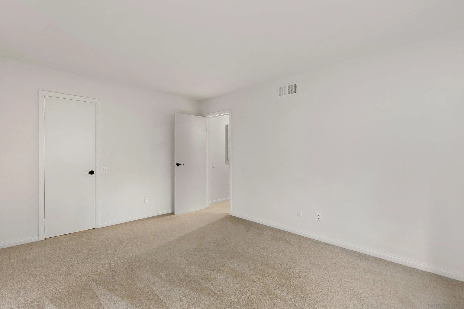 The image showcases a bright, empty bedroom with neutral beige carpeting and white walls, creating a clean and spacious feel. Two white doors are visible, one leading to a closet and the other to an adjacent room, enhancing the room's functionality. The room is well-lit, emphasizing its potential as a comfortable and versatile space.