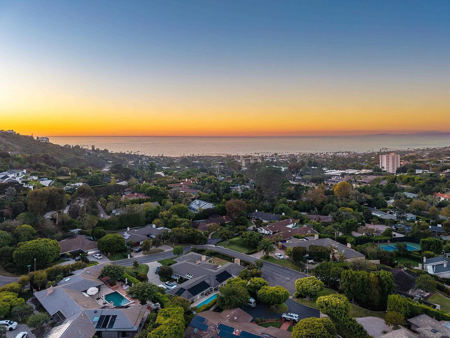 An aerial view showcases a luxurious neighborhood with lush greenery, multiple houses with pools, and meticulously landscaped yards. The scene transitions to the ocean horizon under a vibrant sunset sky, creating a sense of tranquility and upscale living. Solar panels are visible on some roofs, indicating modern eco-consciousness.