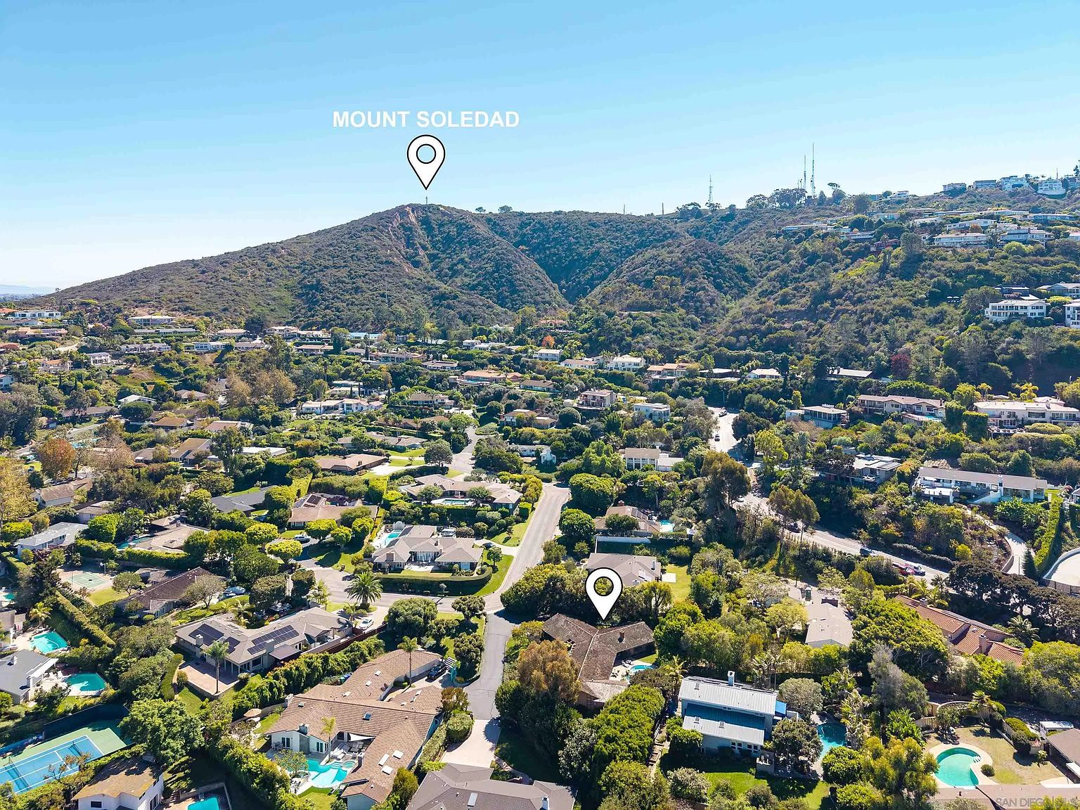 This aerial shot showcases a residential neighborhood nestled in a lush, green landscape with Mount Soledad in the background. The homes feature a variety of architectural styles, many with pools and well-maintained yards, creating an impression of upscale suburban living. The image highlights the proximity to nature and the desirable location.