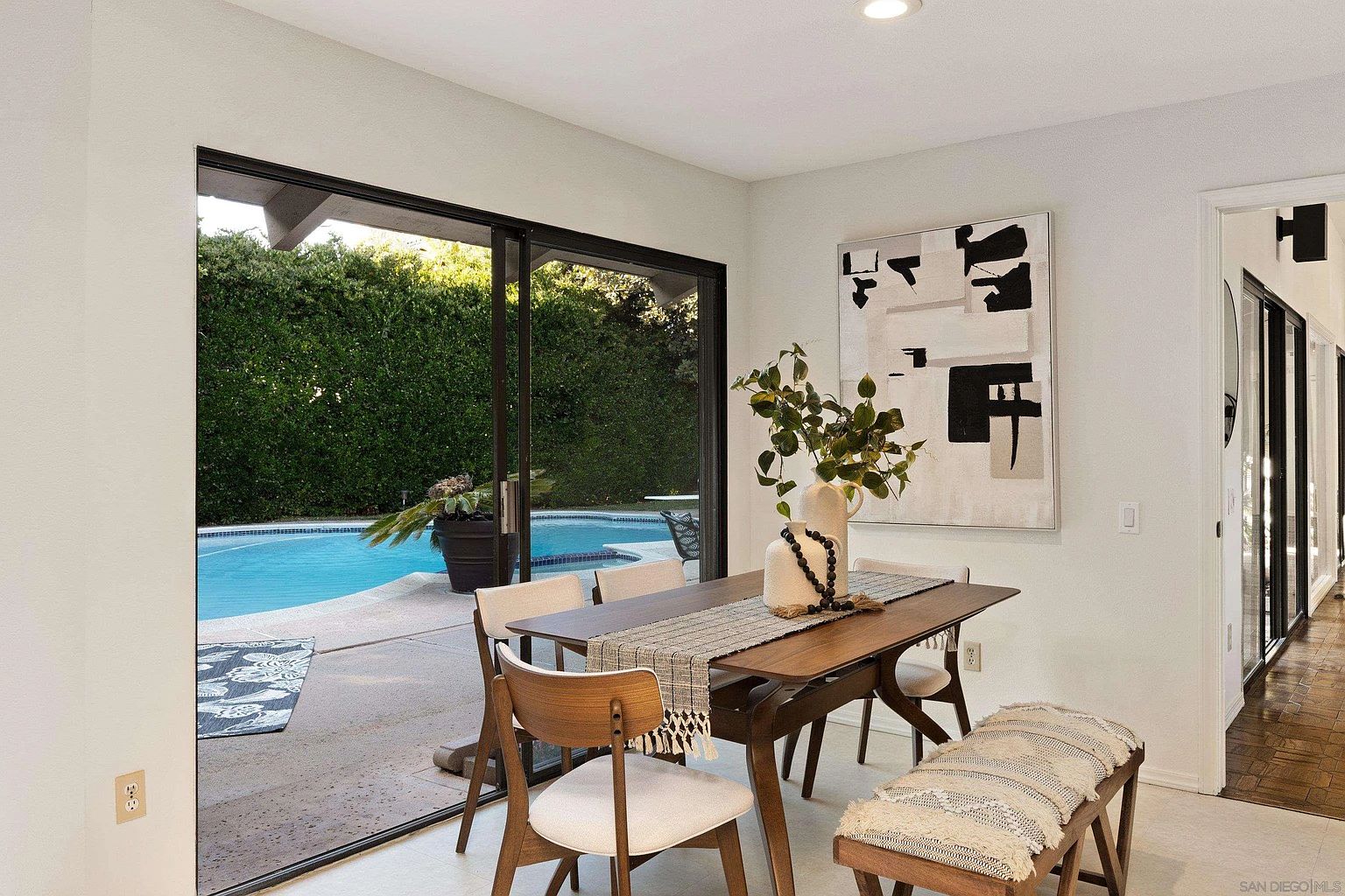 This interior shot showcases a dining room with a modern aesthetic, featuring a wooden dining table set with a decorative runner and vase. The room opens to an outdoor pool area through sliding glass doors, providing a seamless indoor-outdoor living experience. An abstract painting adorns the wall, complementing the room's clean lines and neutral color palette.