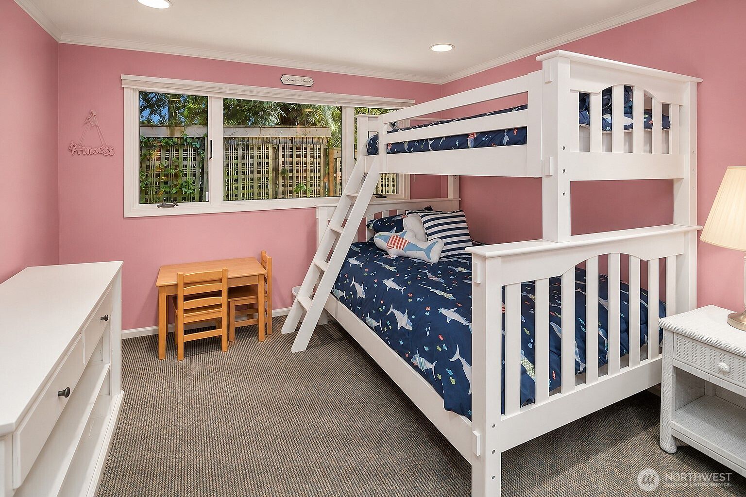 This child's bedroom features soft pink walls and a white bunk bed dressed in a playful shark-patterned quilt. A small wooden table and chair set sits near the window, which looks out onto a fenced outdoor area. The room is completed by a white dresser and a matching nightstand, creating a cozy and functional space.