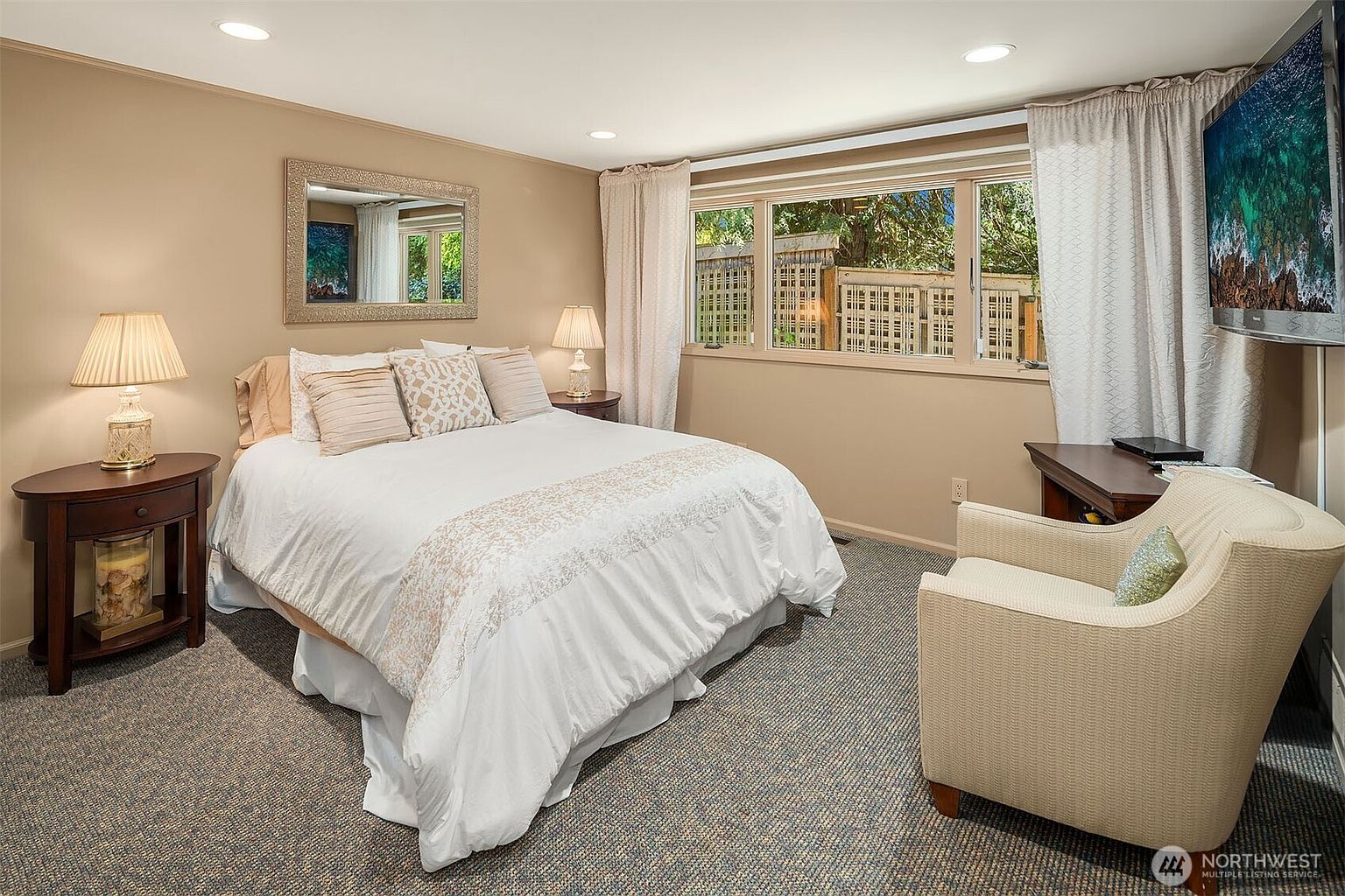 This cozy bedroom features a queen-sized bed centered against a neutral-toned wall, flanked by two dark wood nightstands with matching table lamps. A large window with light curtains allows natural light to brighten the space, while a comfortable armchair sits in the foreground near a desk. The room is finished with neutral carpeting and a decorative mirror hanging above the headboard, creating a warm and inviting atmosphere.
