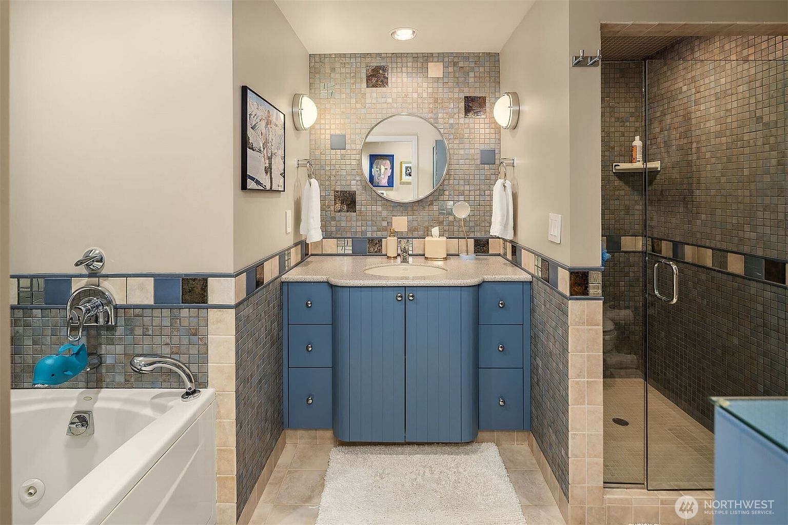 This bathroom features a blue vanity with a light-colored countertop, centered beneath a round mirror and flanked by wall-mounted light fixtures. To the left, a soaking tub is equipped with a chrome faucet, while a glass-enclosed shower stall with tiled walls sits to the right. The room is accented with a decorative mosaic tile backsplash and a neutral-toned tiled floor, creating a cohesive and functional space.