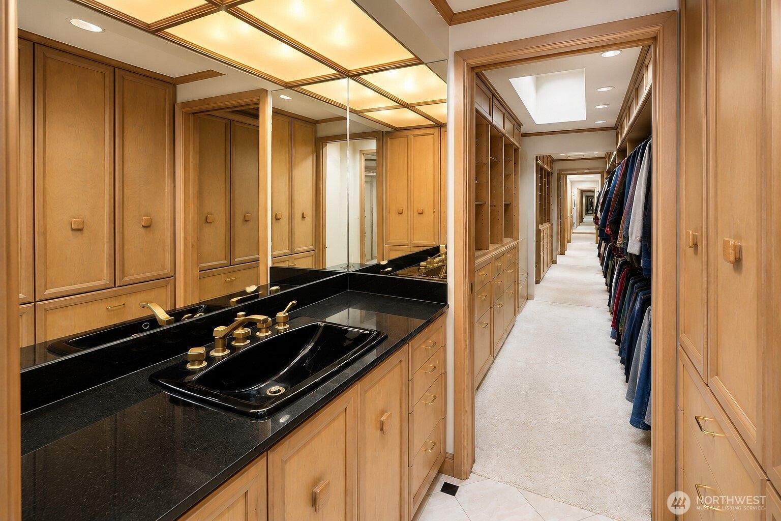 This primary bathroom features a vanity with a dark stone countertop and a black sink, complemented by light wood cabinetry and gold-toned fixtures. A large mirror spans the wall above the vanity, reflecting the adjacent walk-in closet which is lined with extensive wooden shelving and hanging space. The perspective looks from the vanity area into the long, carpeted closet, creating a seamless transition between the two functional spaces.
